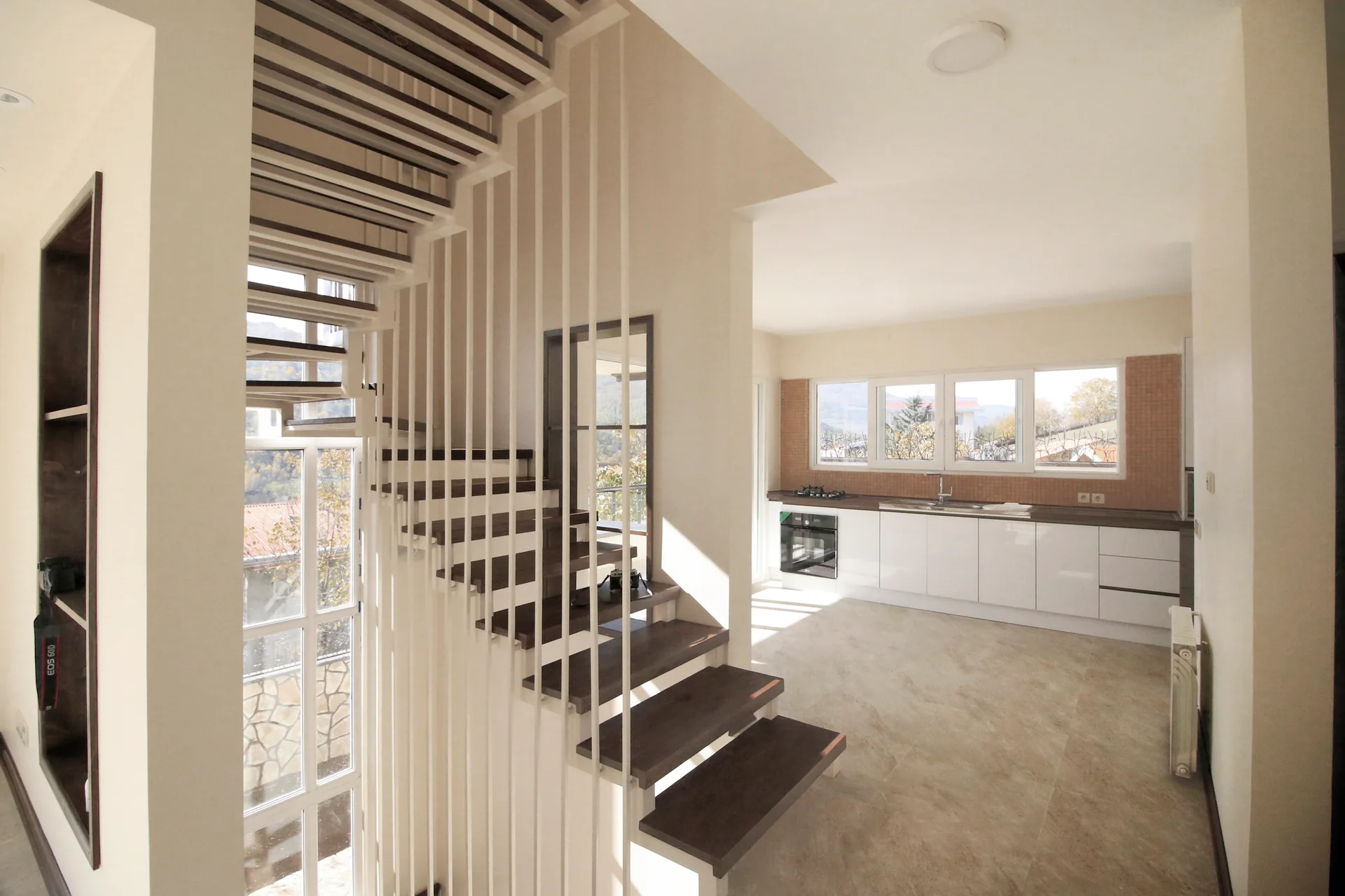 A modern open-plan interior featuring a floating wooden staircase with white vertical rails alongside a minimalist white kitchen with tiled backsplash.