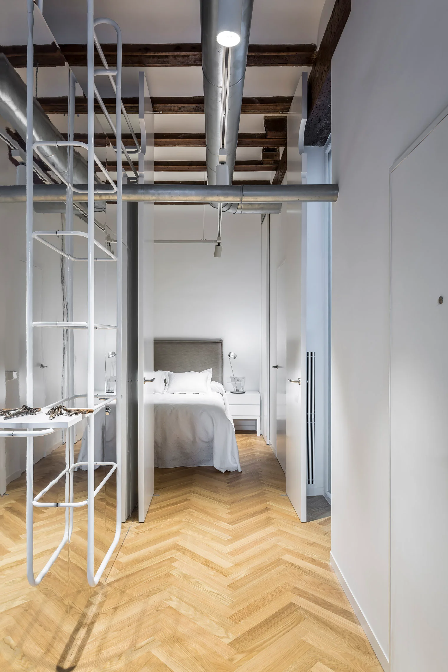 A modern bedroom framed by white walls features a bed, exposed ceiling beams, industrial metal ductwork, and herringbone wood floors.