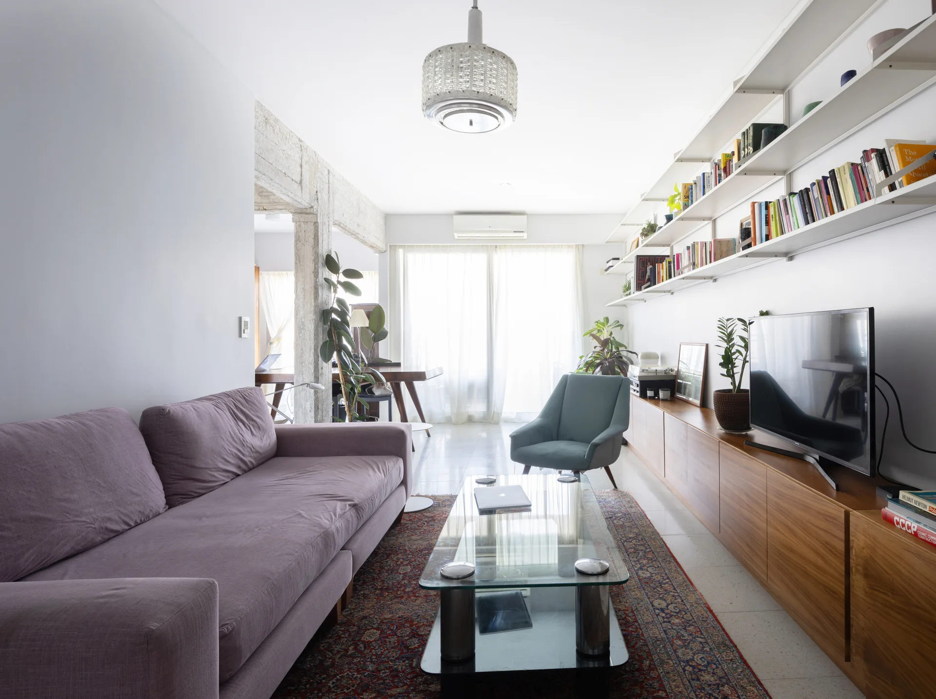 A bright living room features a purple sofa, a glass coffee table, a long wooden media unit, and multiple white floating shelves filled with books.