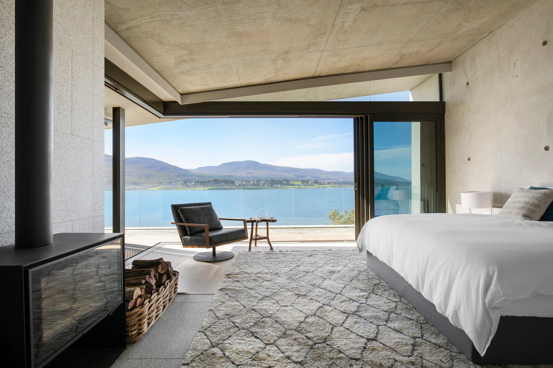 A minimalist bedroom featuring raw concrete walls, a large bed with white linens, and sliding glass doors that look out over a scenic lake.