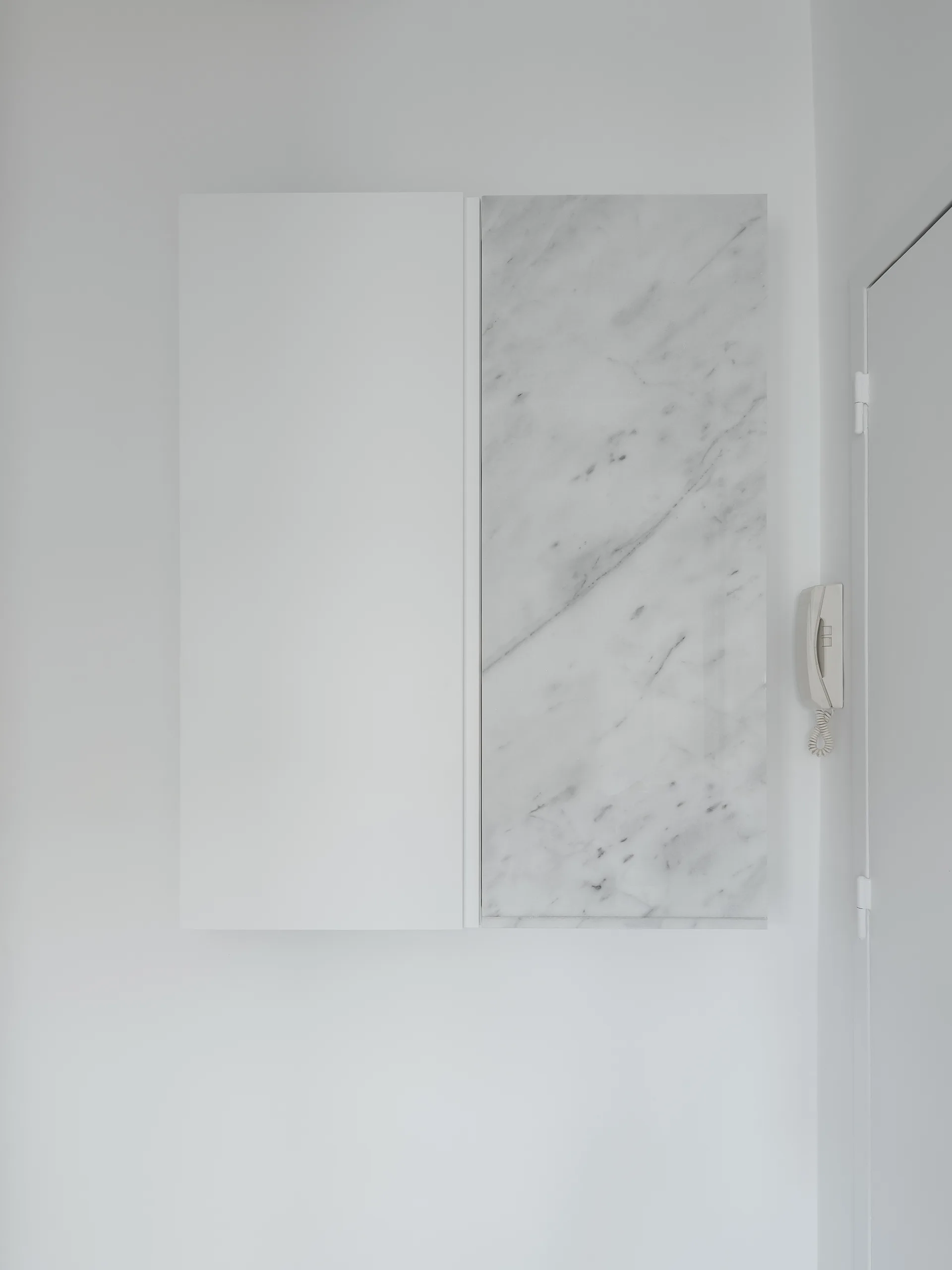 A minimalist wall-mounted cabinet with one matte white panel and one veined white marble panel hangs on a plain white wall next to an intercom.