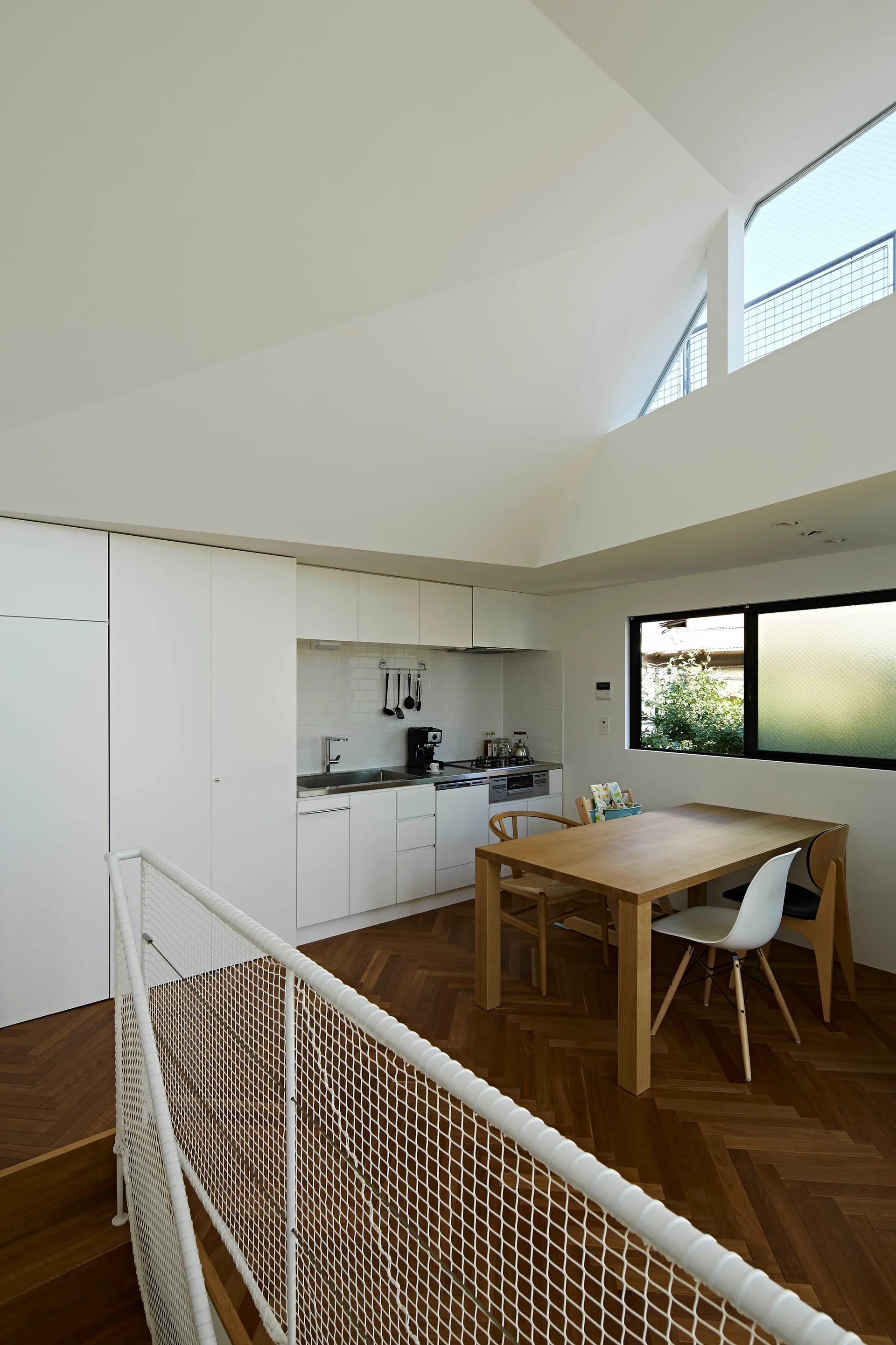 A modern white kitchen and dining area features a wooden table, herringbone wood floors, and minimalist cabinets under a high vaulted ceiling.