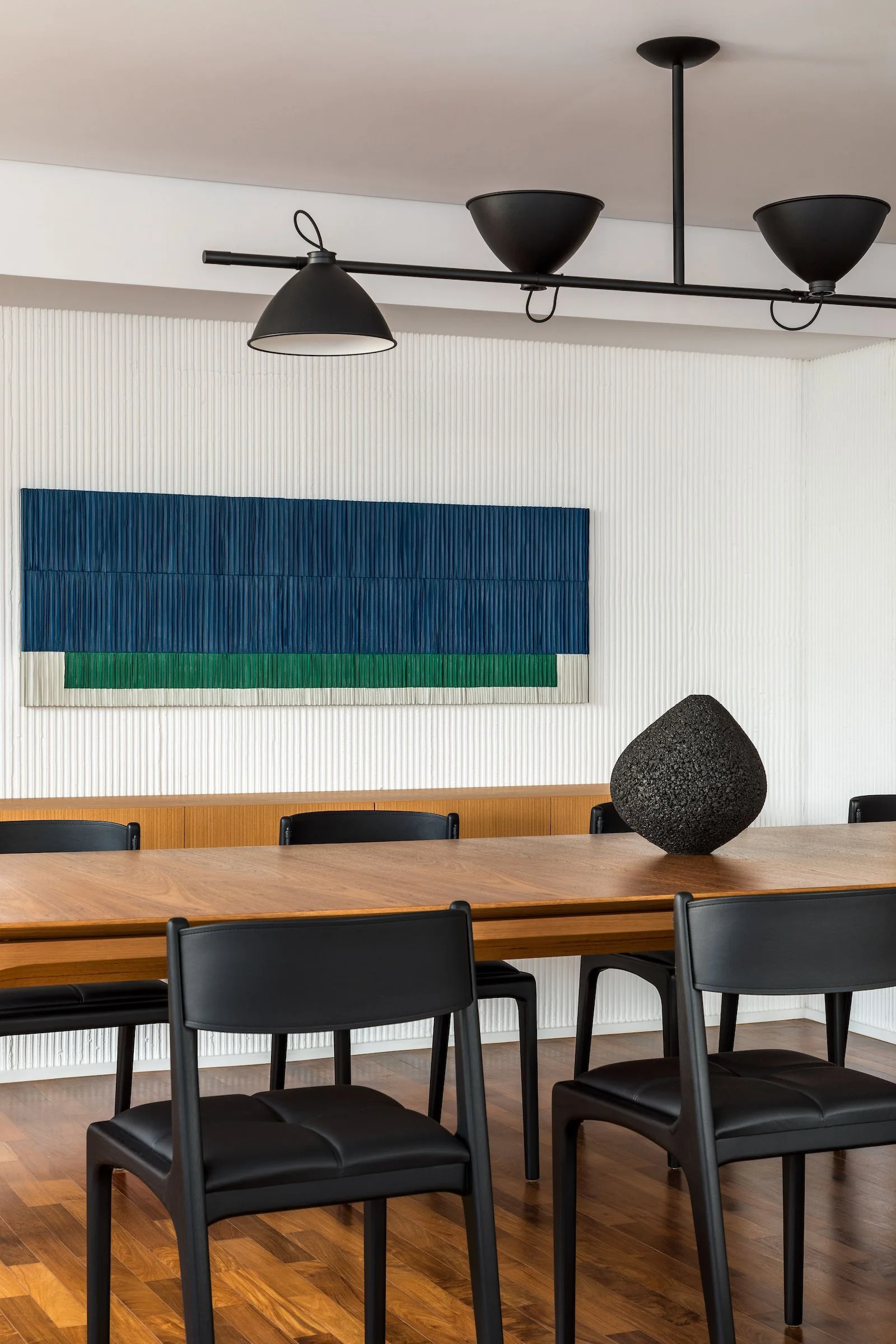 A wooden dining table surrounded by black leather chairs sits in front of a white fluted accent wall displaying a blue textured artwork.