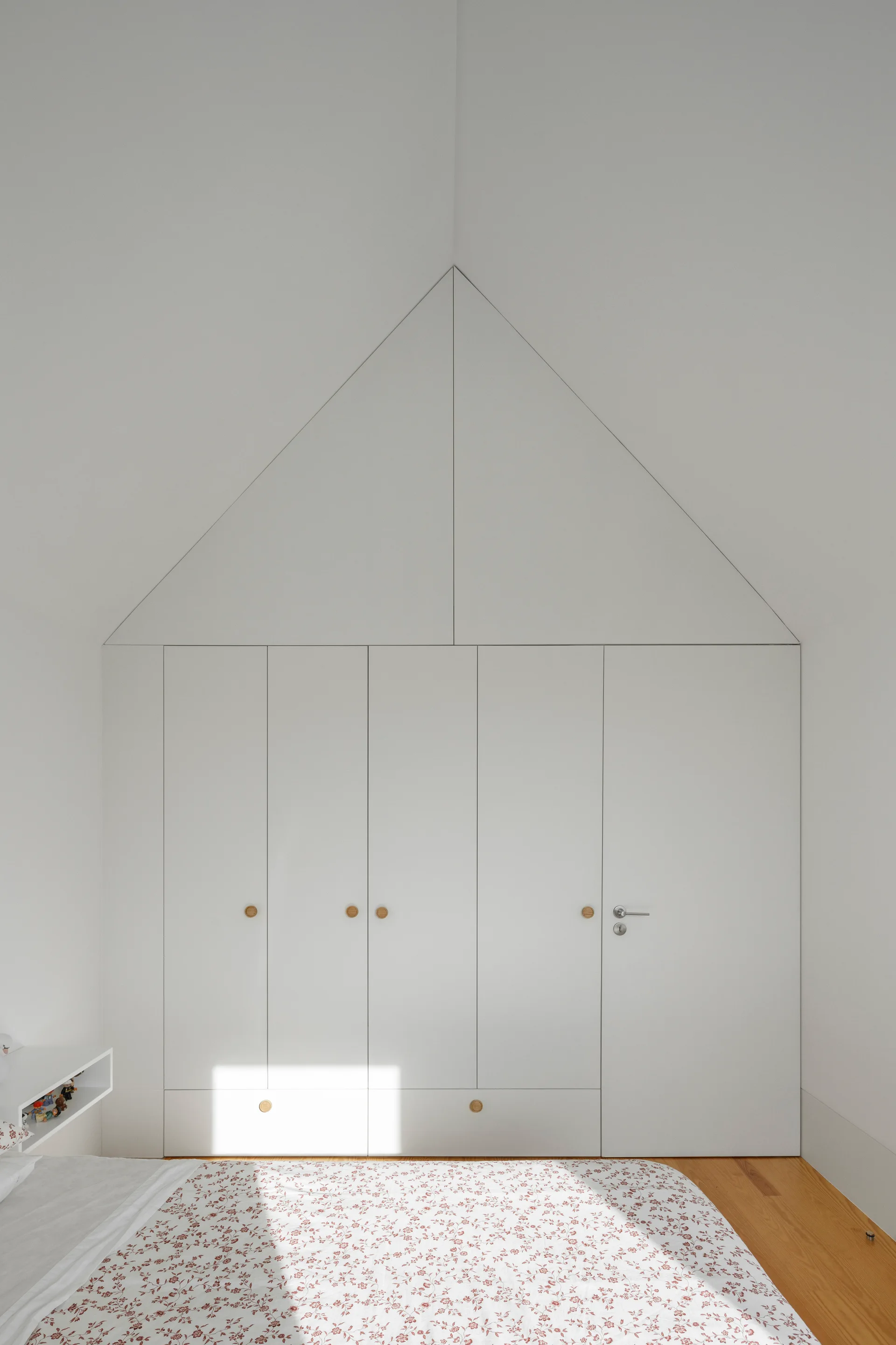 A minimalist white built-in wardrobe features a triangular house-shaped top panel, round wooden knobs, and integrated lower drawers in a bedroom.