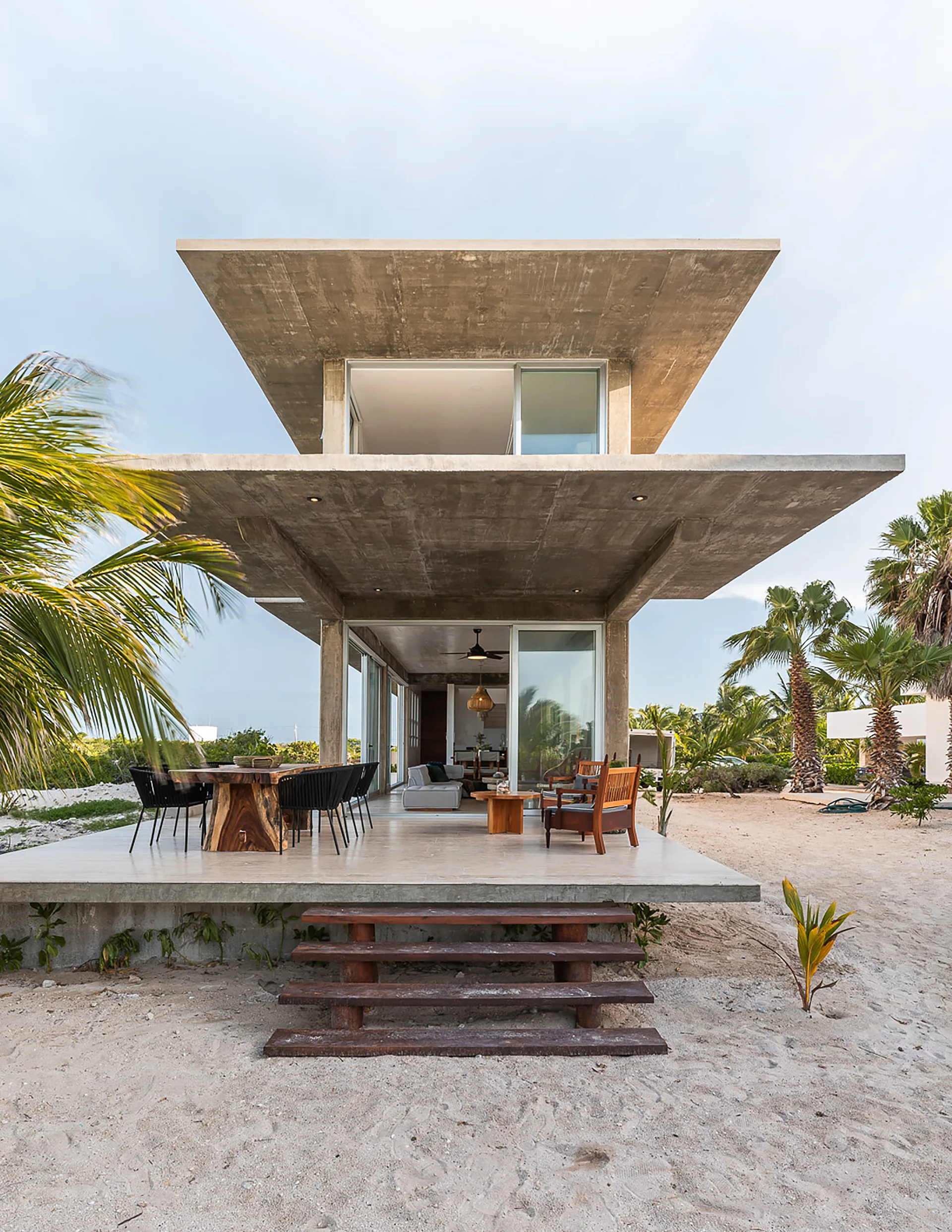A modern concrete beach house features a raised patio with a live-edge wooden dining table, black chairs, and steps leading to the sand.