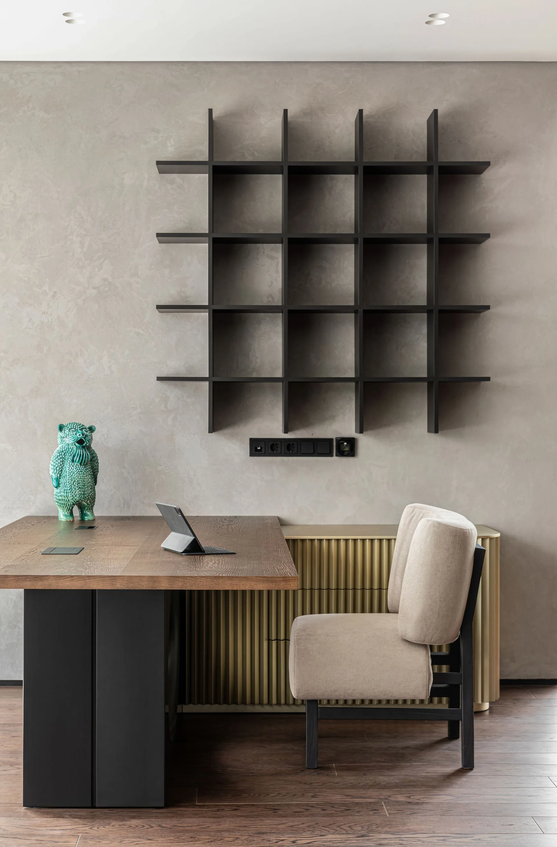 A contemporary home office features a dark wood desk, a beige upholstered chair, a gold fluted cabinet, and a black grid shelf on a textured grey wall.