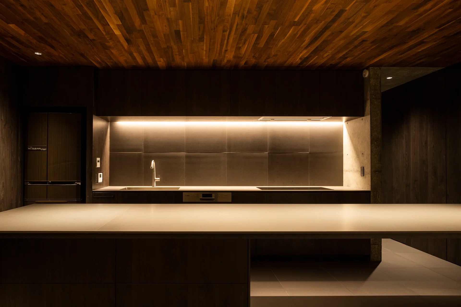 A modern kitchen featuring a warm wood slat ceiling, dark wood cabinets with integrated lighting, and a smooth stone island.