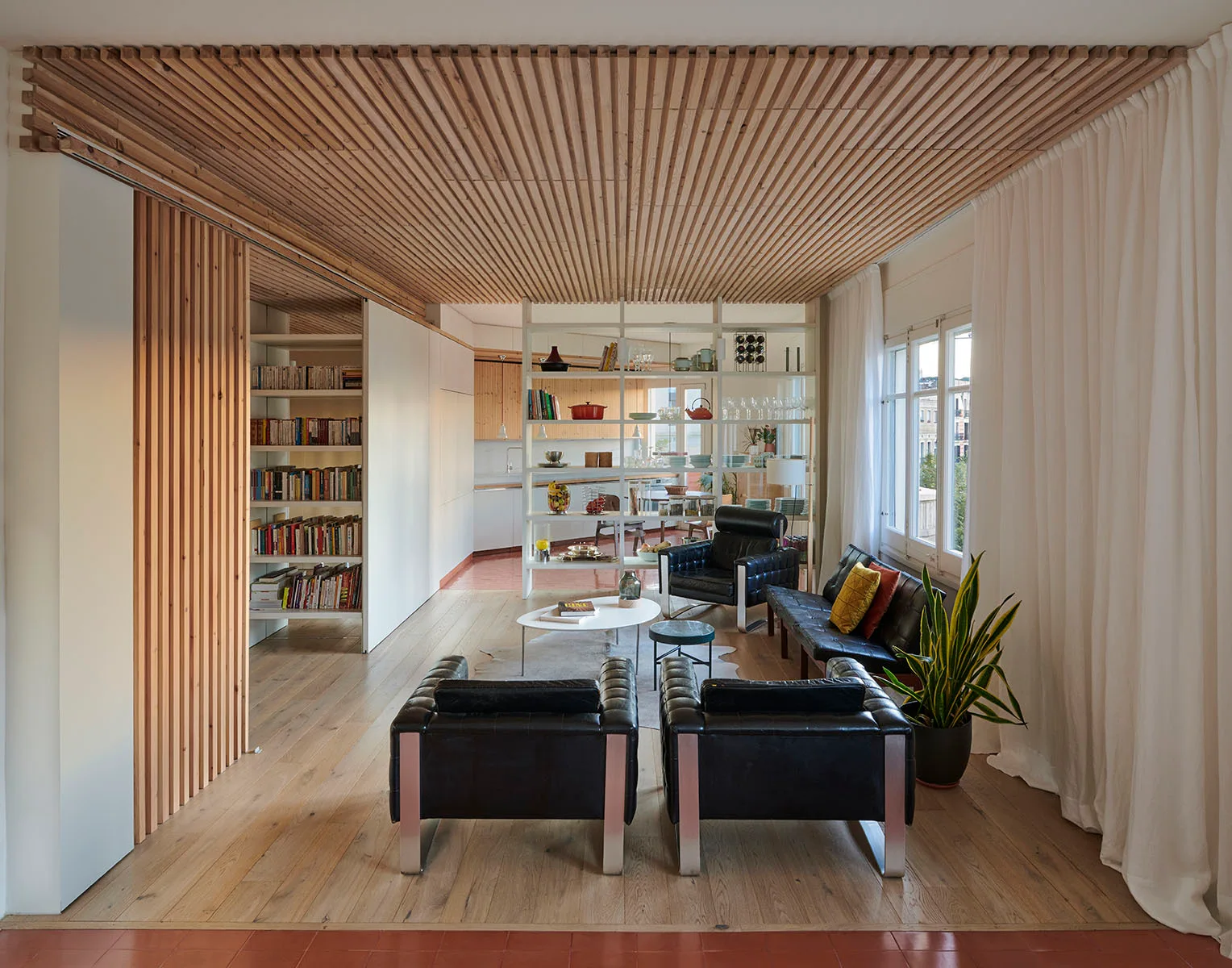 A modern living room features a slatted wood ceiling, black leather armchairs, and an open shelving unit acting as a room divider.