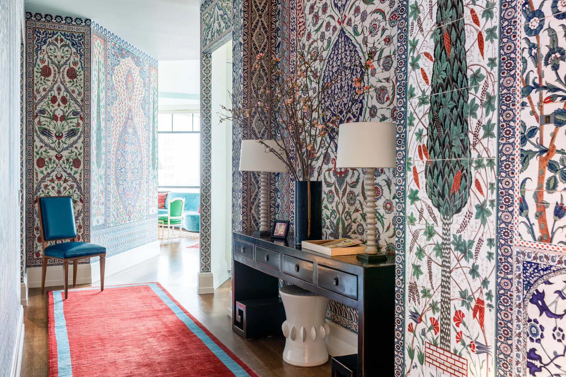 A maximalist hallway features intricate floral ceramic tile walls, a dark wood console table with lamps, and a red runner rug.