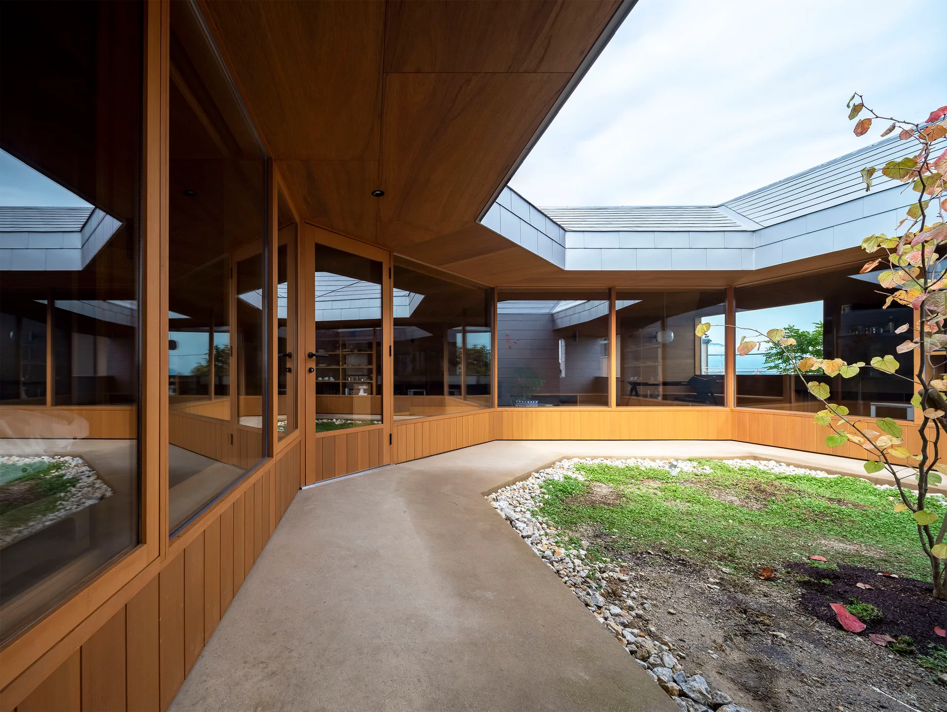 A concrete path curves around a small central garden inside a modern courtyard surrounded by wood-trimmed glass walls and a matching wooden soffit.