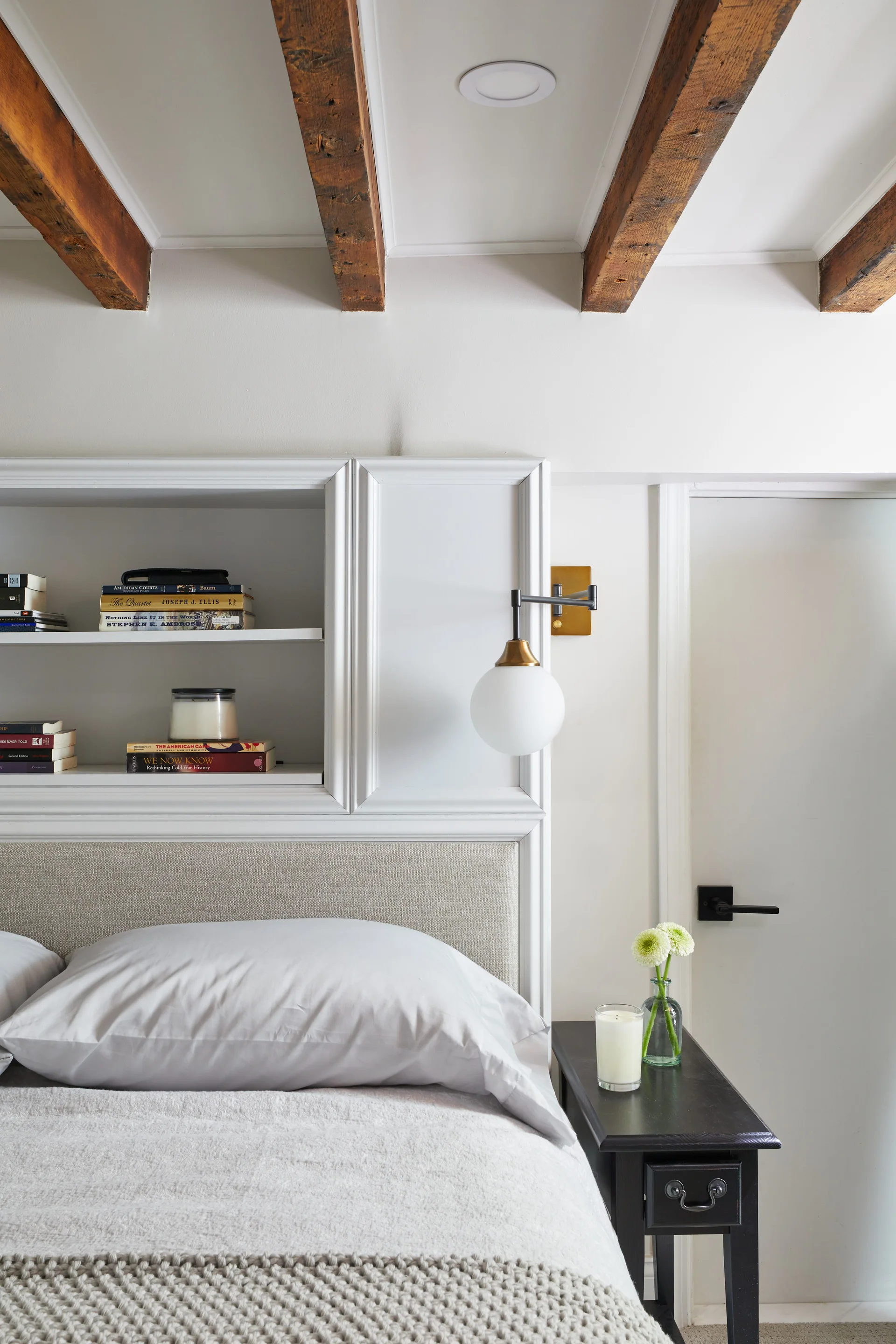 A white upholstered headboard built into a custom shelving unit beneath rustic wood ceiling beams.