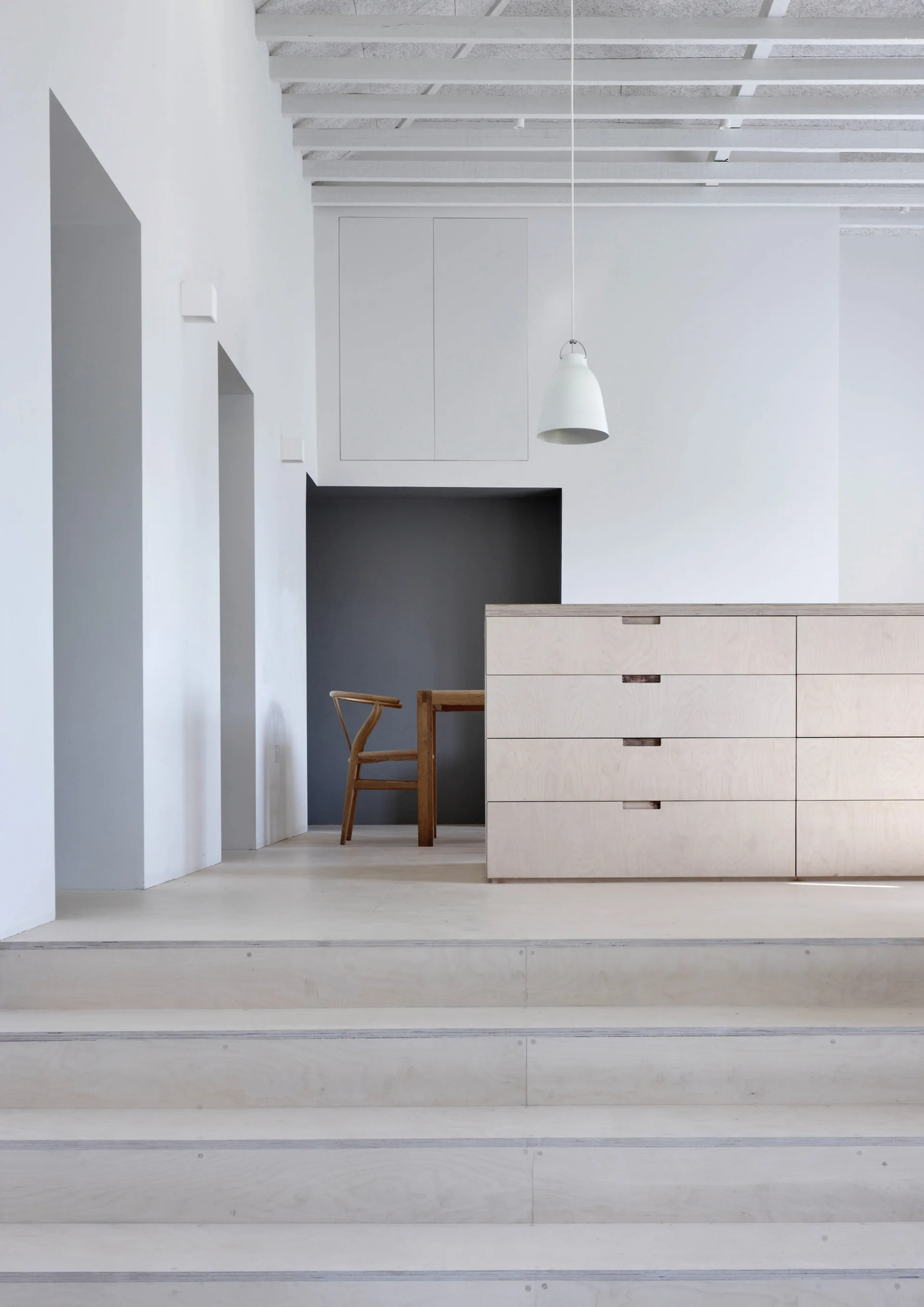 Plywood stairs lead up to a minimalist kitchen featuring a light wood drawer unit, white walls, ceiling joists, and a dining chair in a grey recess.