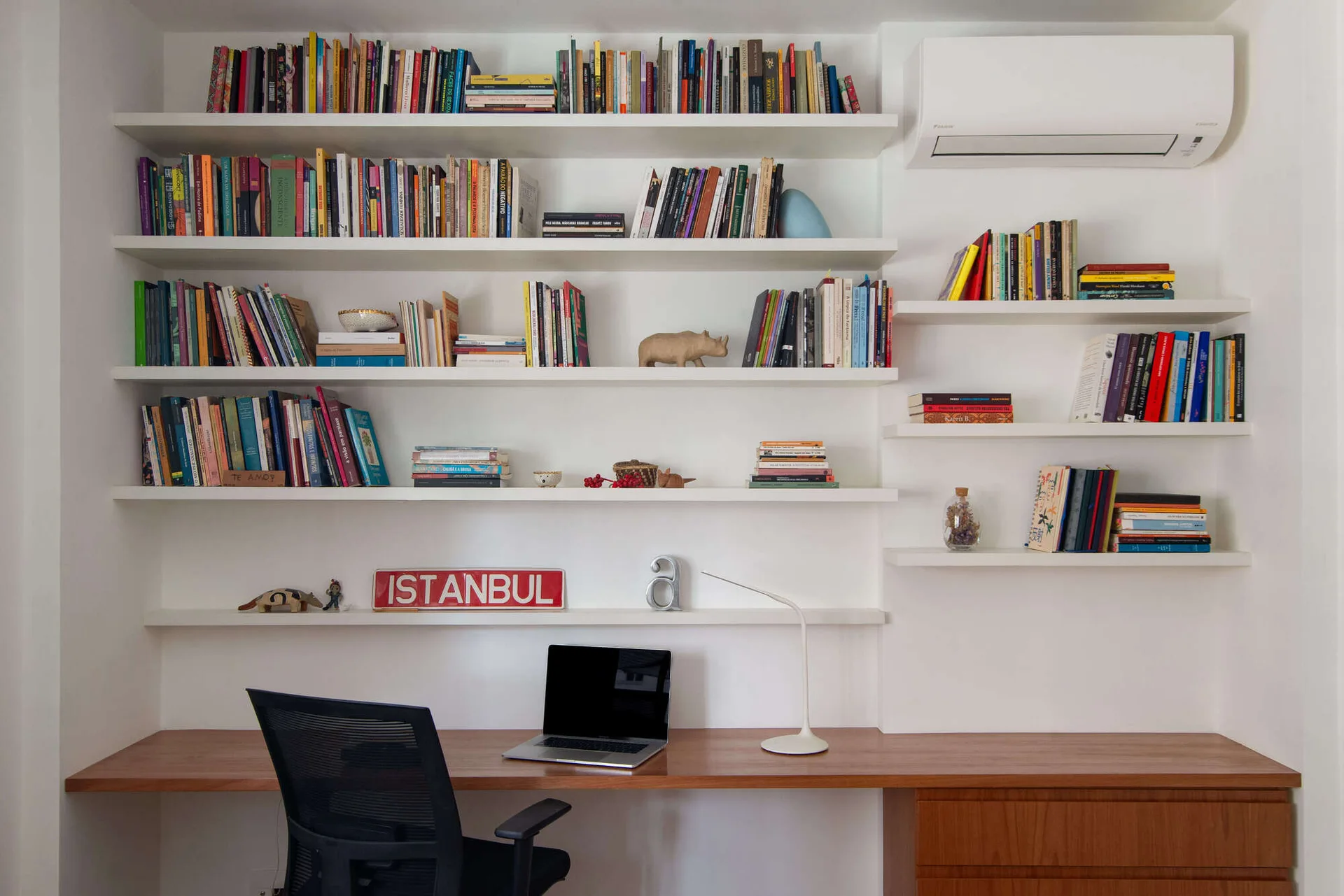 A modern home office features a long wooden desk, a black mesh chair, and white wall-mounted shelves filled with books.