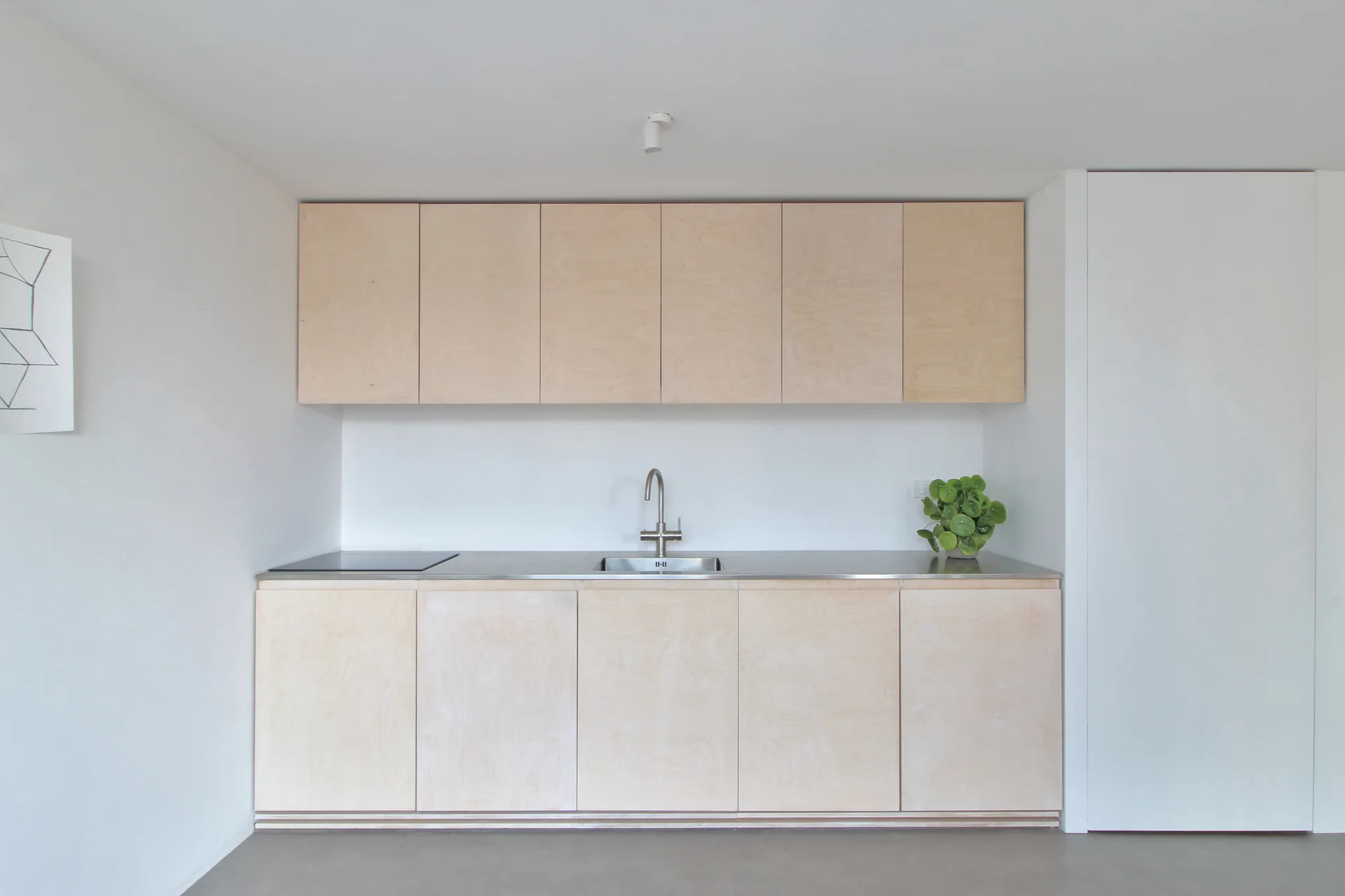 A minimalist kitchen featuring light wood plywood cabinets, a integrated stainless steel countertop and sink, and white walls.