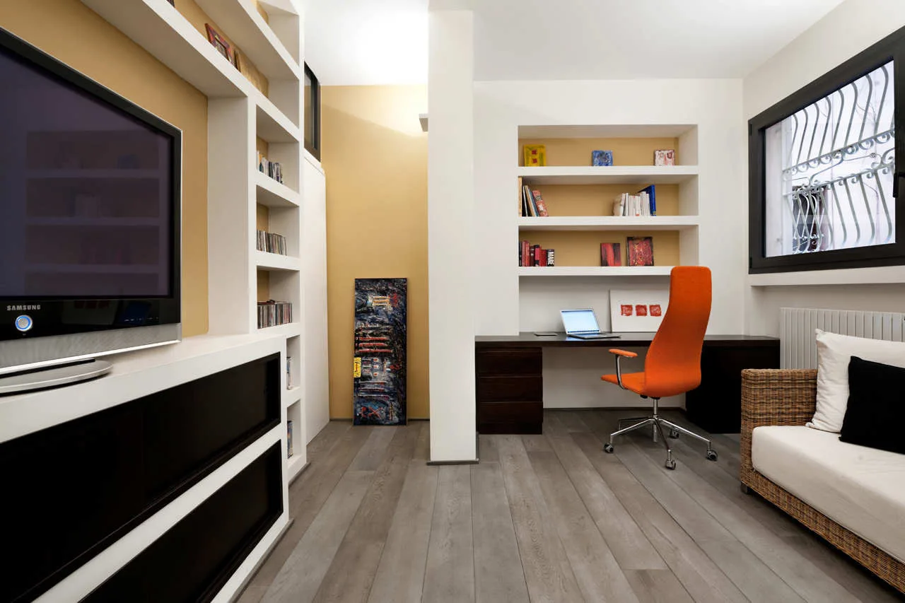 A home office features a dark wood desk with an orange swivel chair, white built-in wall shelves with gold backings, and grey wood floors.