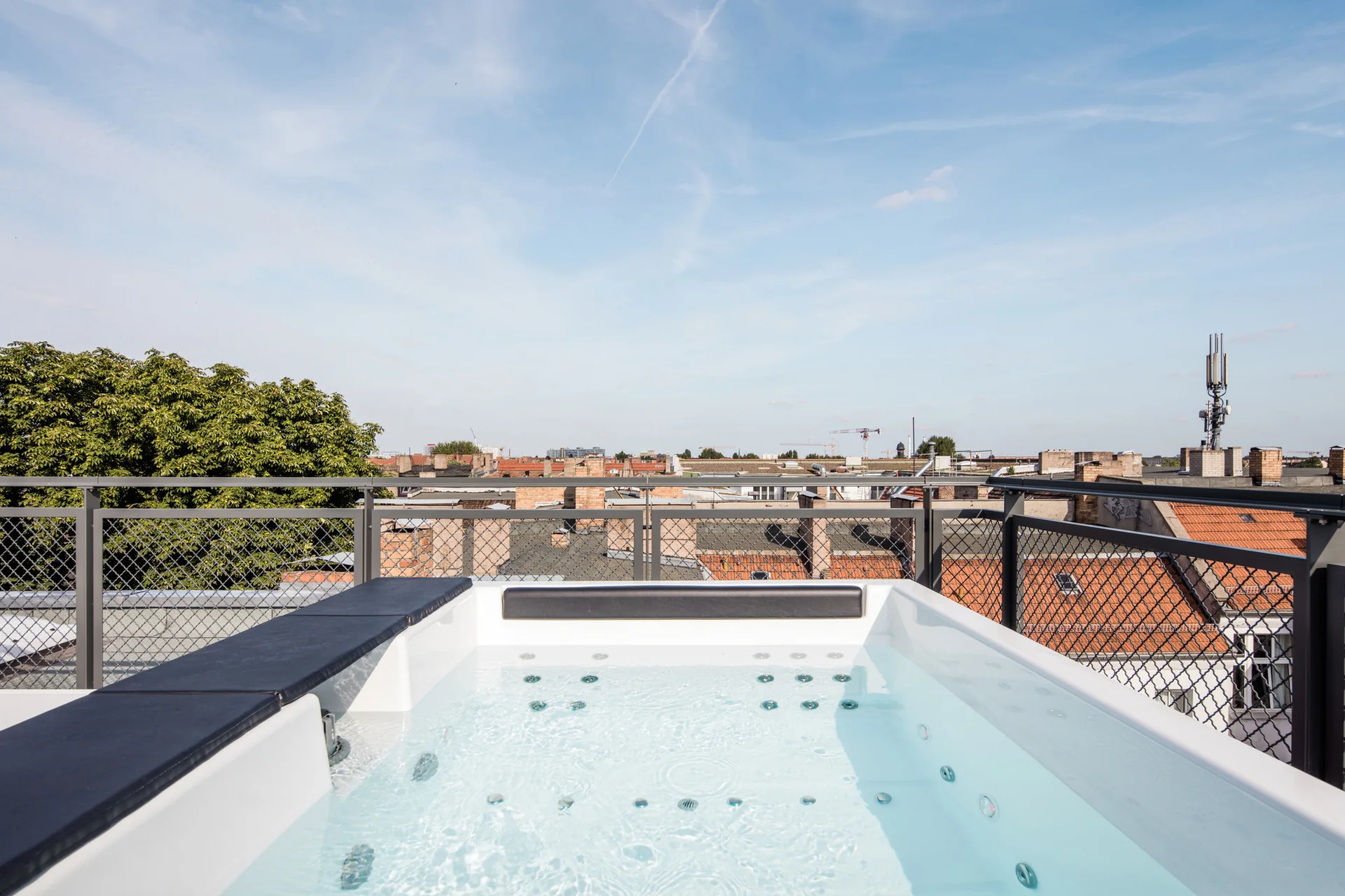 A white hot tub filled with water on a rooftop terrace, enclosed by a black metal mesh railing overlooking a city under a clear blue sky.