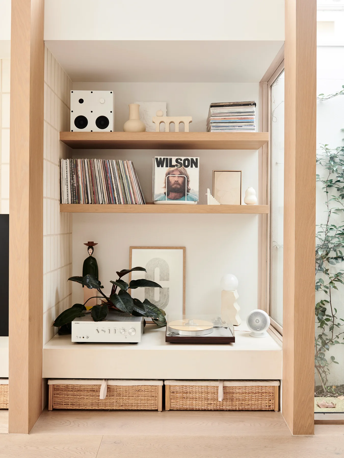 A light oak built-in shelving unit displays a vinyl record player, audio amplifier, speakers, ceramic art, and wicker storage baskets.