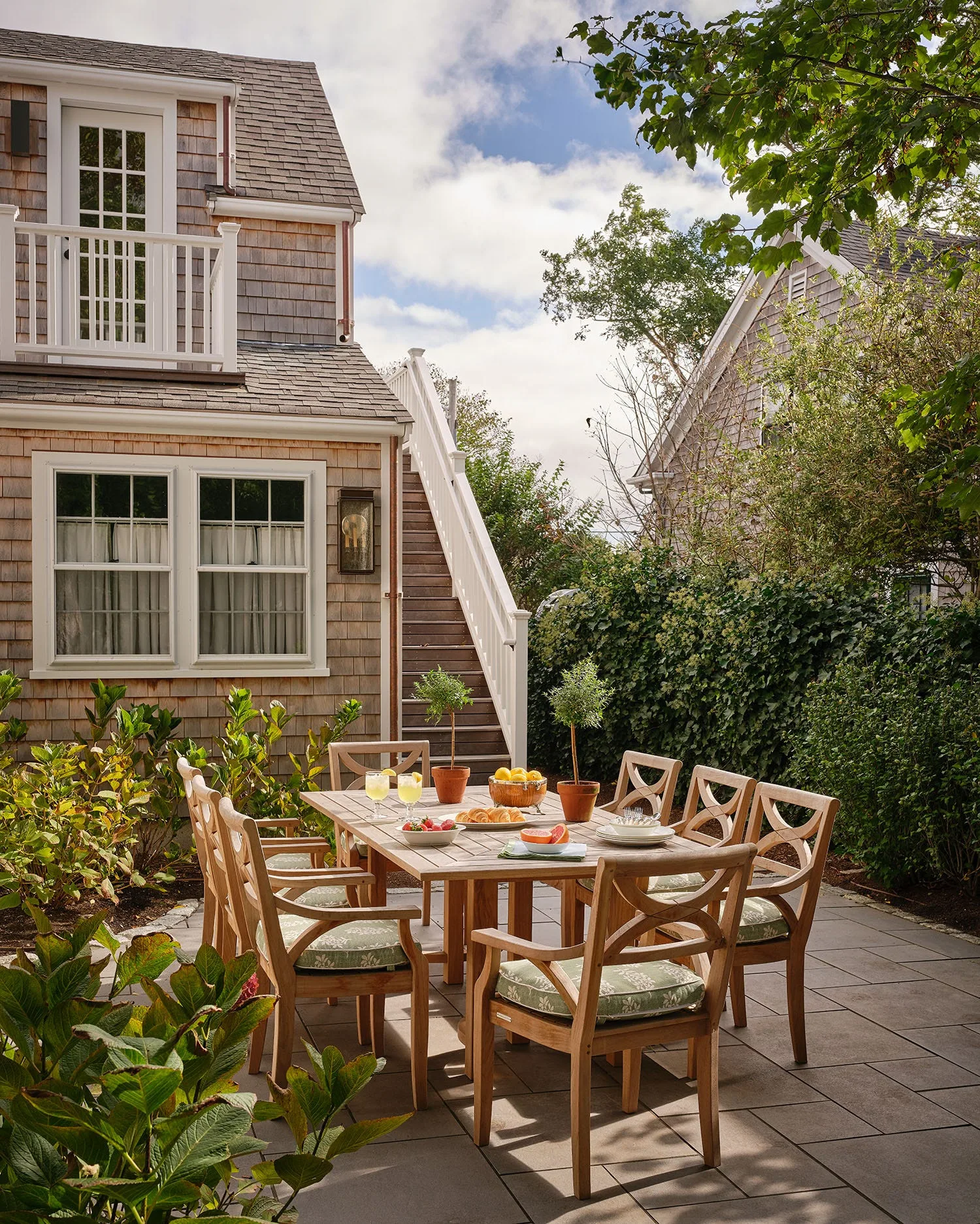 A wooden dining table and chairs with green patterned cushions are set on a grey stone patio next to a shingle-sided house.