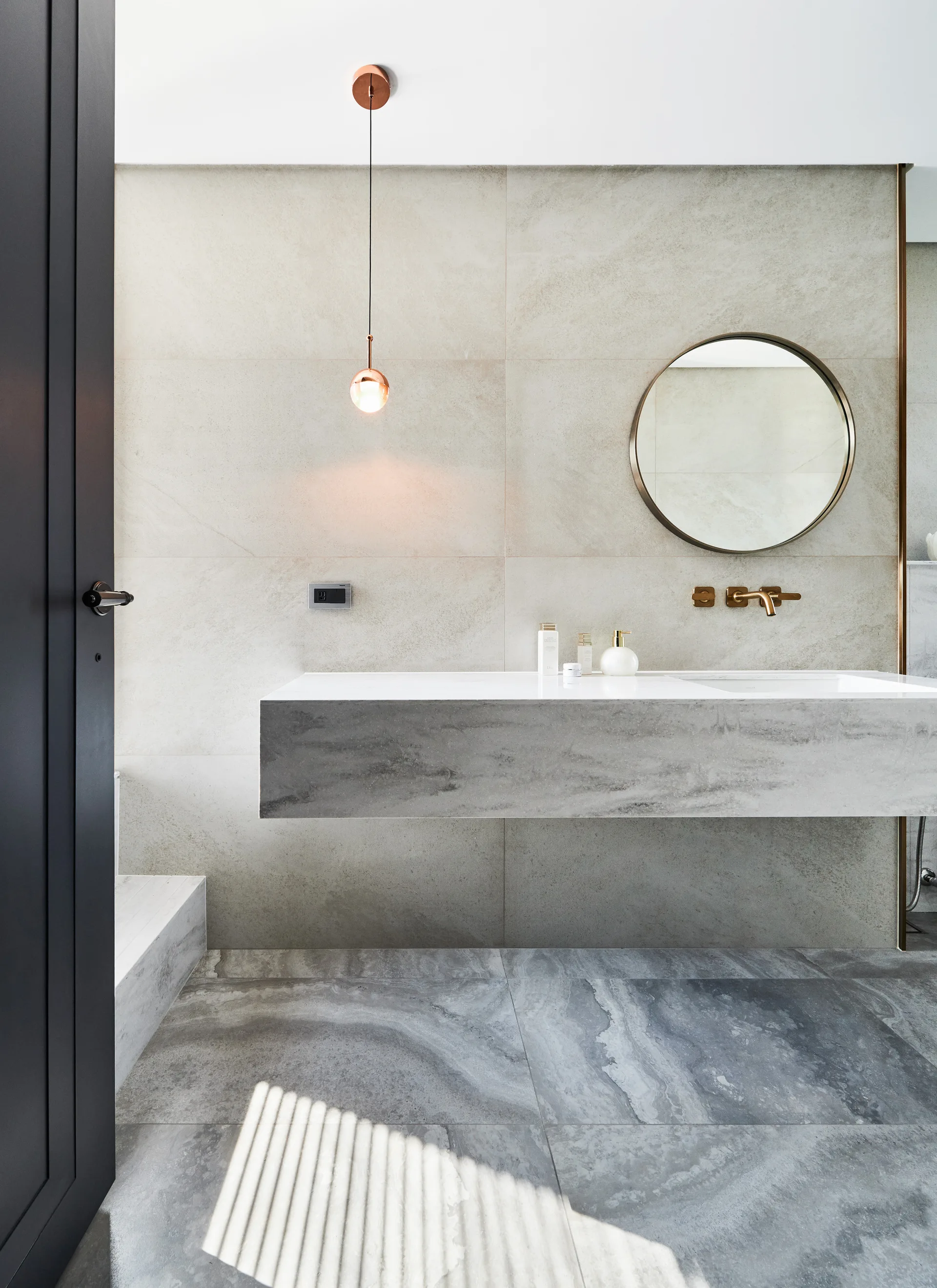 A modern bathroom features a floating stone vanity, a wall-mounted gold faucet, a round mirror, and a hanging copper pendant light.