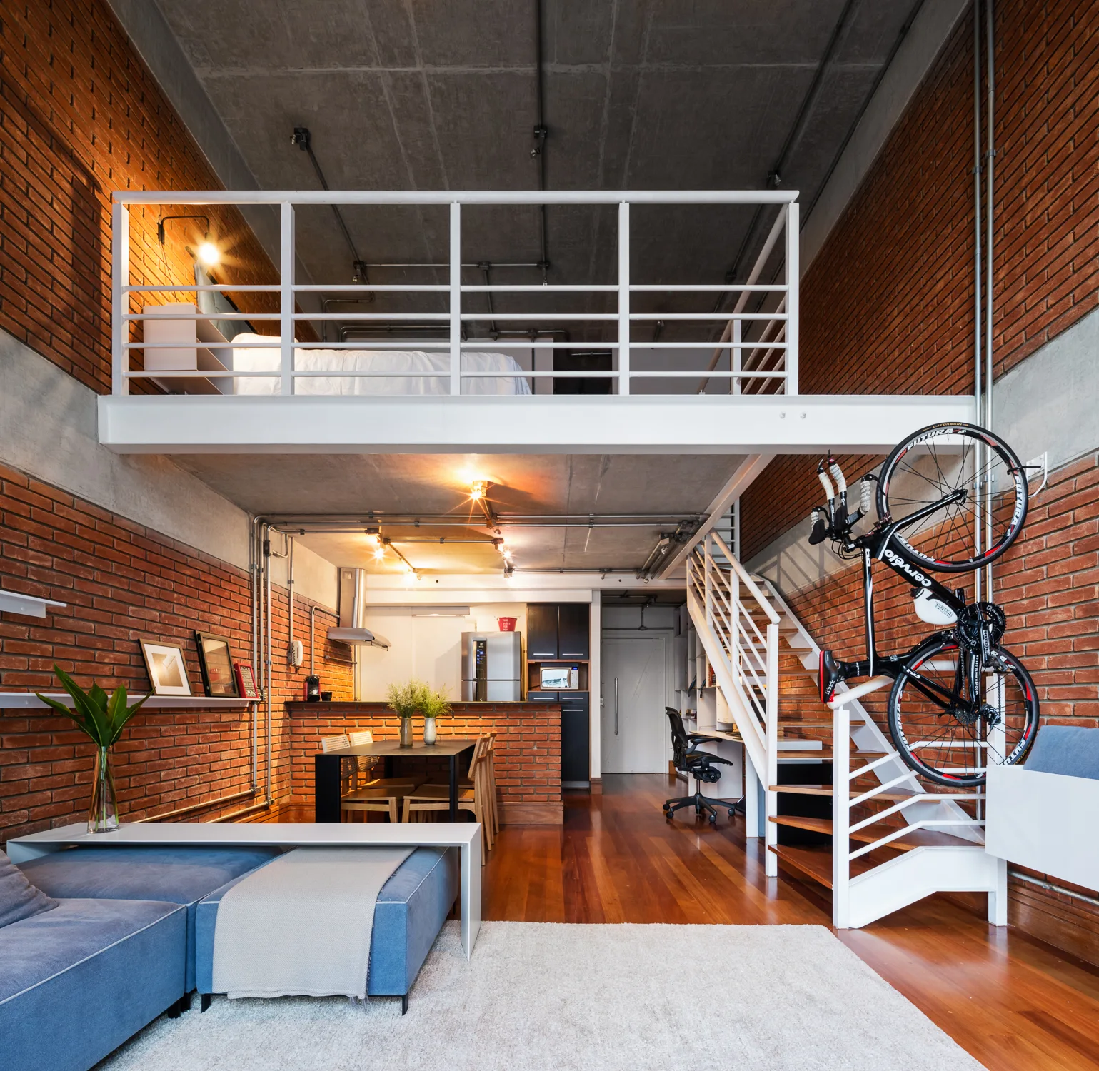 An industrial loft apartment with red brick walls, a concrete ceiling, a white metal mezzanine bedroom, a staircase, and a blue sofa.