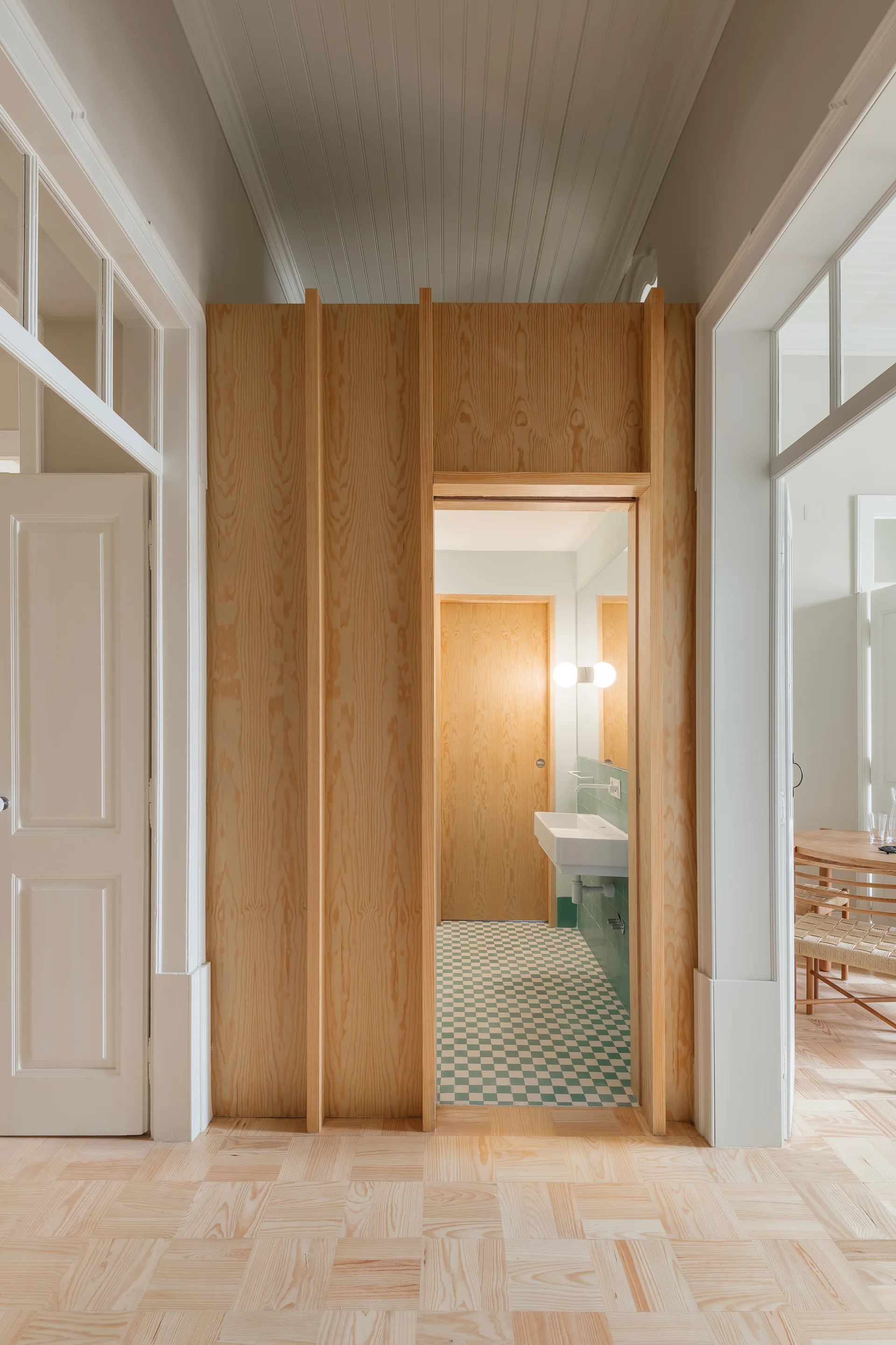 A light wood paneled hallway looks into a small bathroom featuring green and white checkerboard floor tiles and a wall-mounted sink.