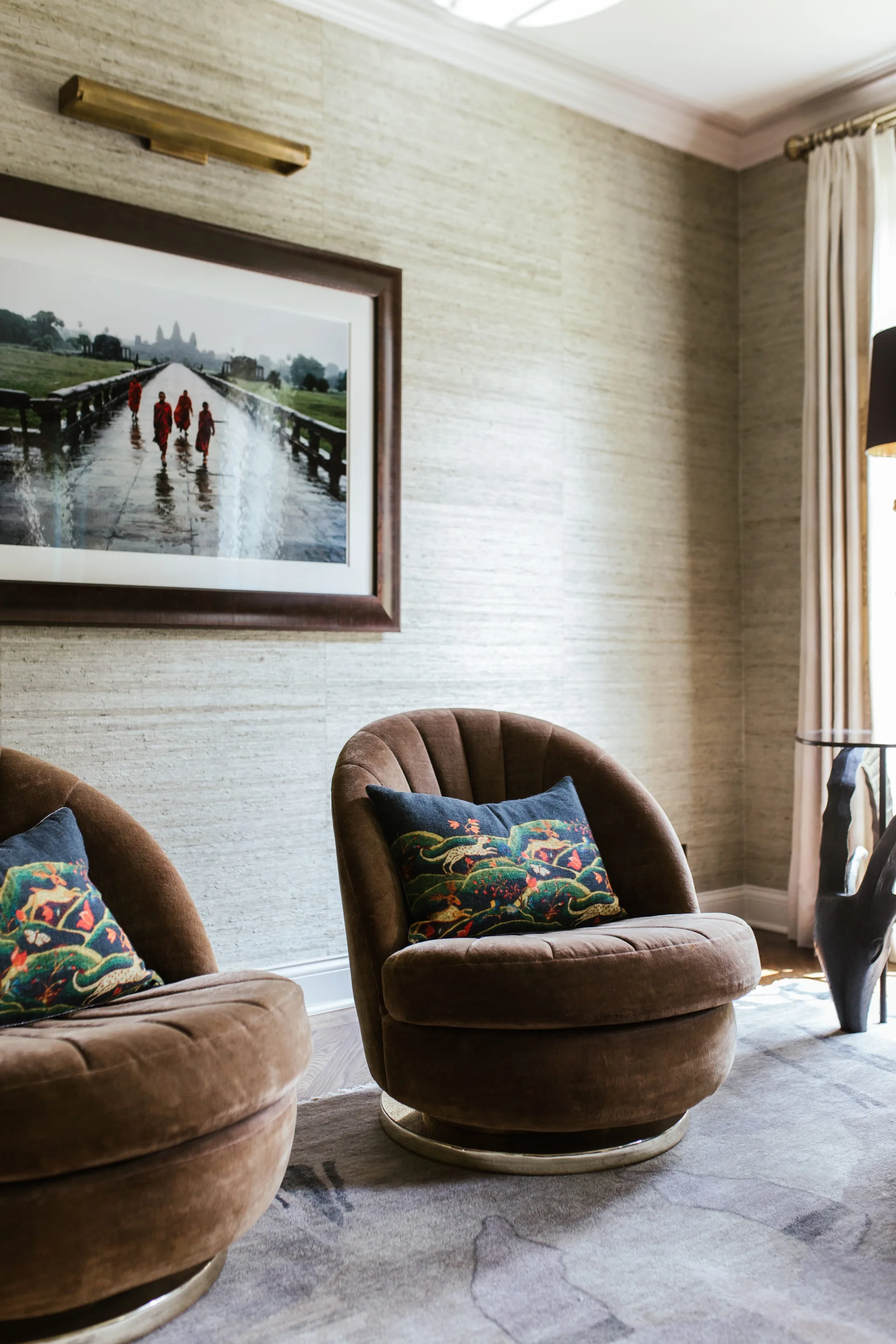Two brown velvet swivel armchairs with patterned pillows sit on an abstract patterned rug against a textured grasscloth wall with framed art.