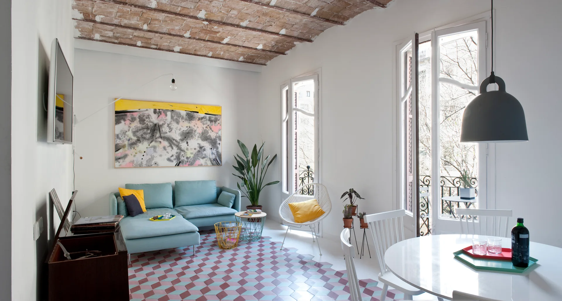 An open space features a light blue sectional sofa, multi-colored geometric floor tiles, and an exposed brick vaulted ceiling.