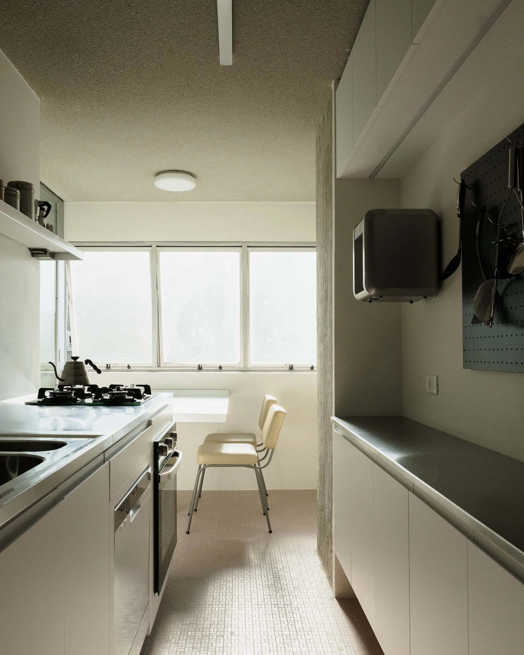 A narrow white galley kitchen features matte cabinetry, a built-in dining table under a wide window, and a wall-mounted utensil pegboard.