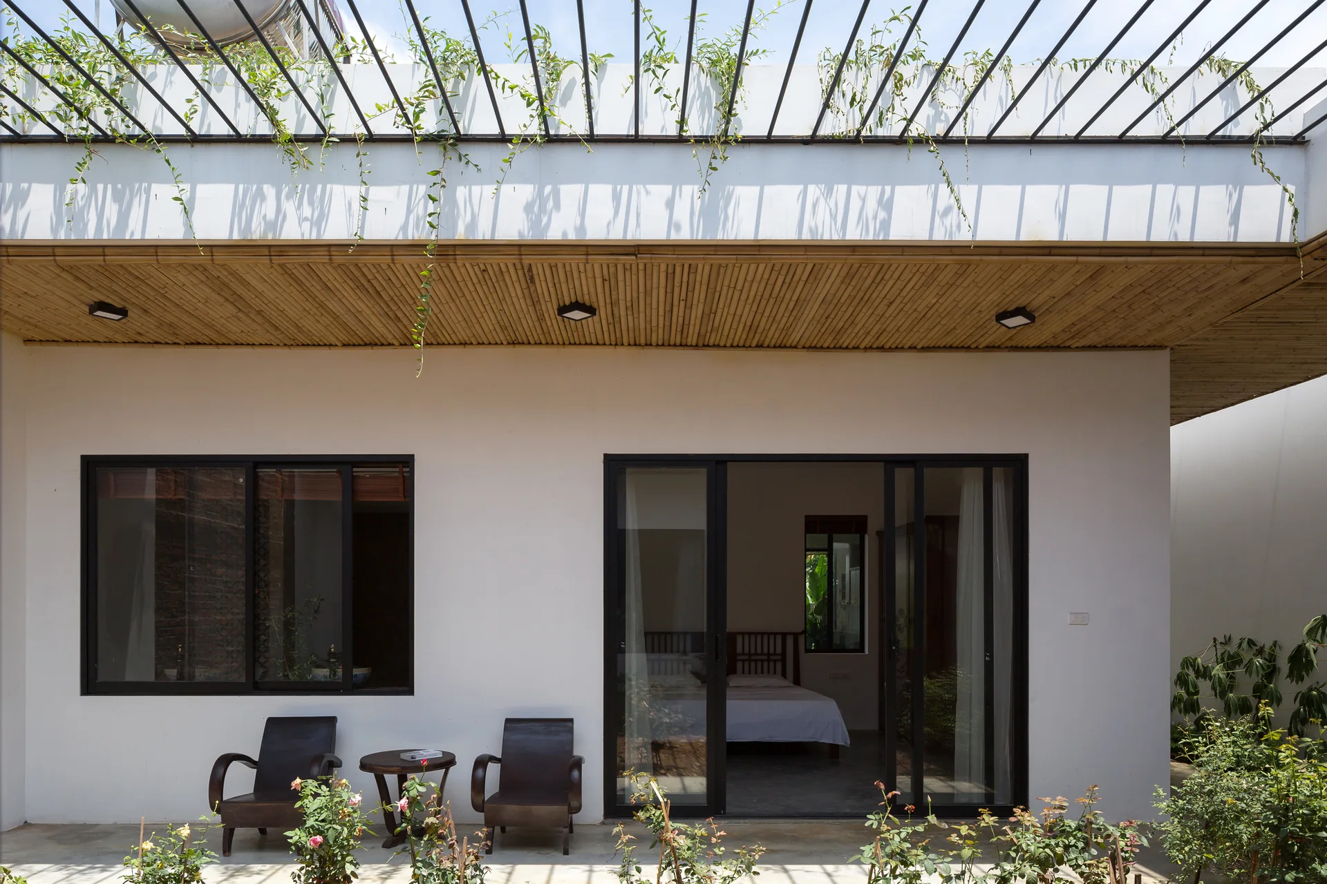 Two dark wood armchairs sit outside on a patio under a slatted bamboo ceiling next to black-framed sliding glass doors.