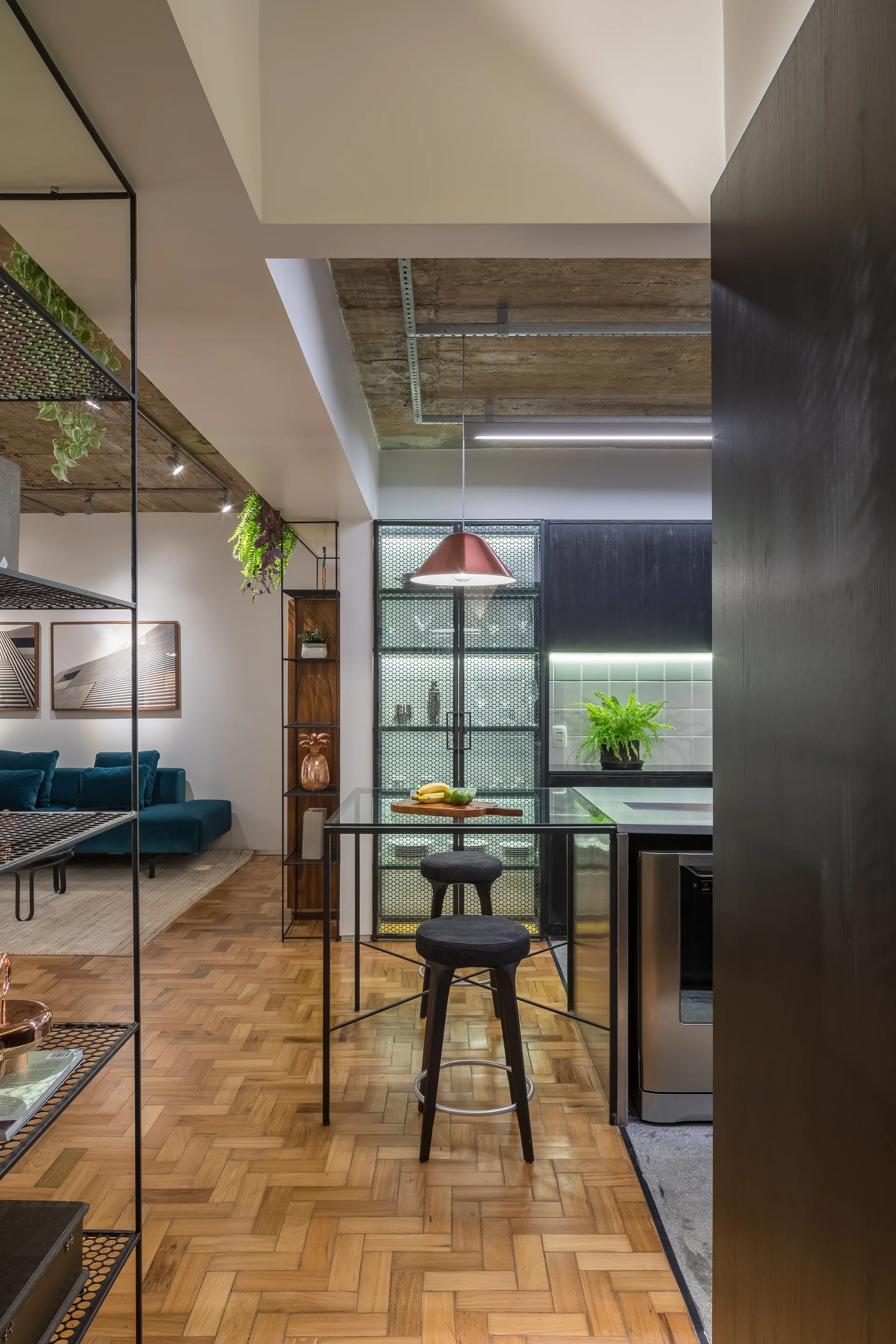 A modern kitchen featuring a glass-topped island with stools, a copper pendant light, herringbone parquet floors, and an exposed concrete ceiling.