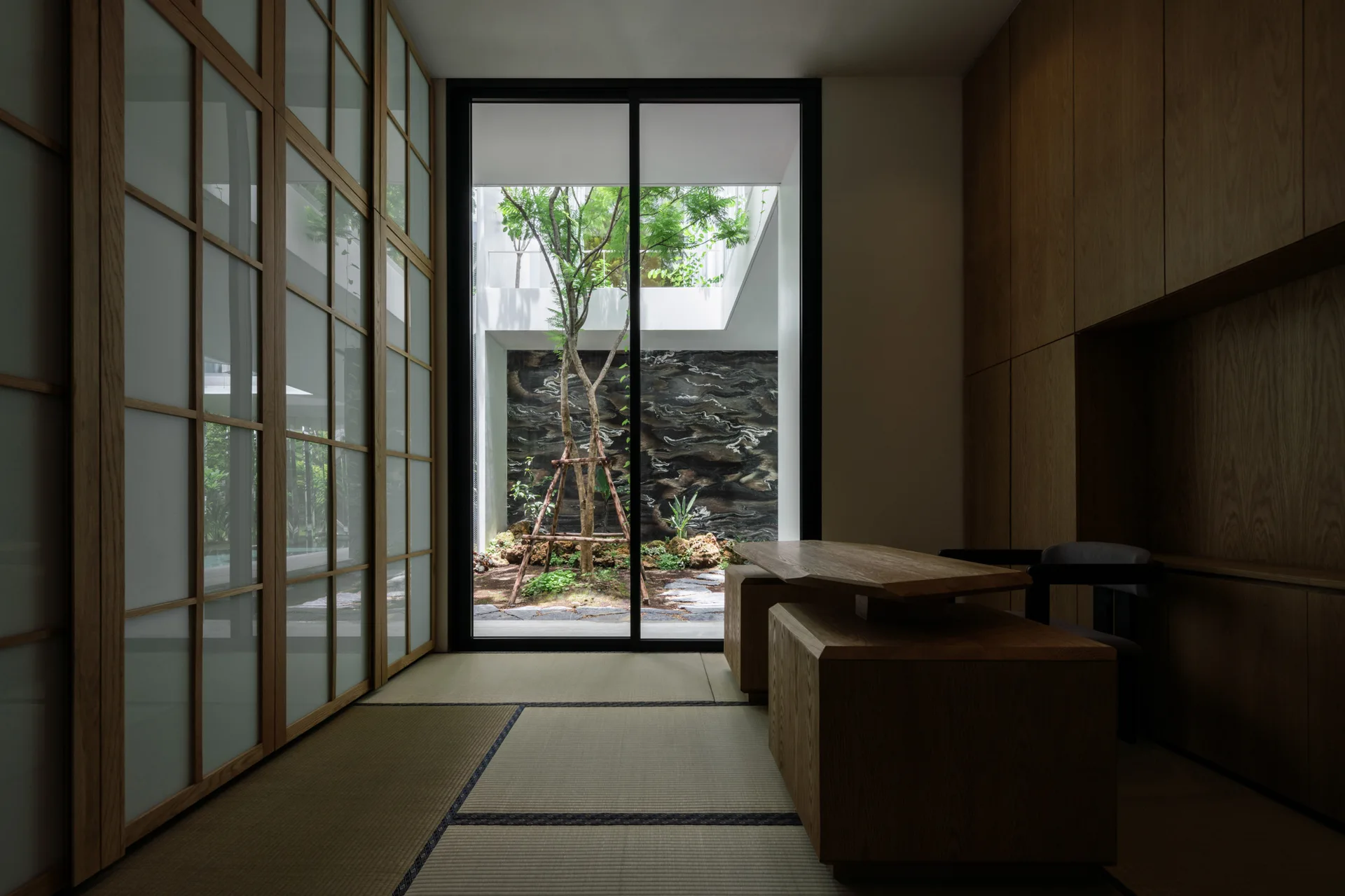 A minimalist study features tatami mat flooring, a low wooden desk, and a large glass door looking out to a garden courtyard.