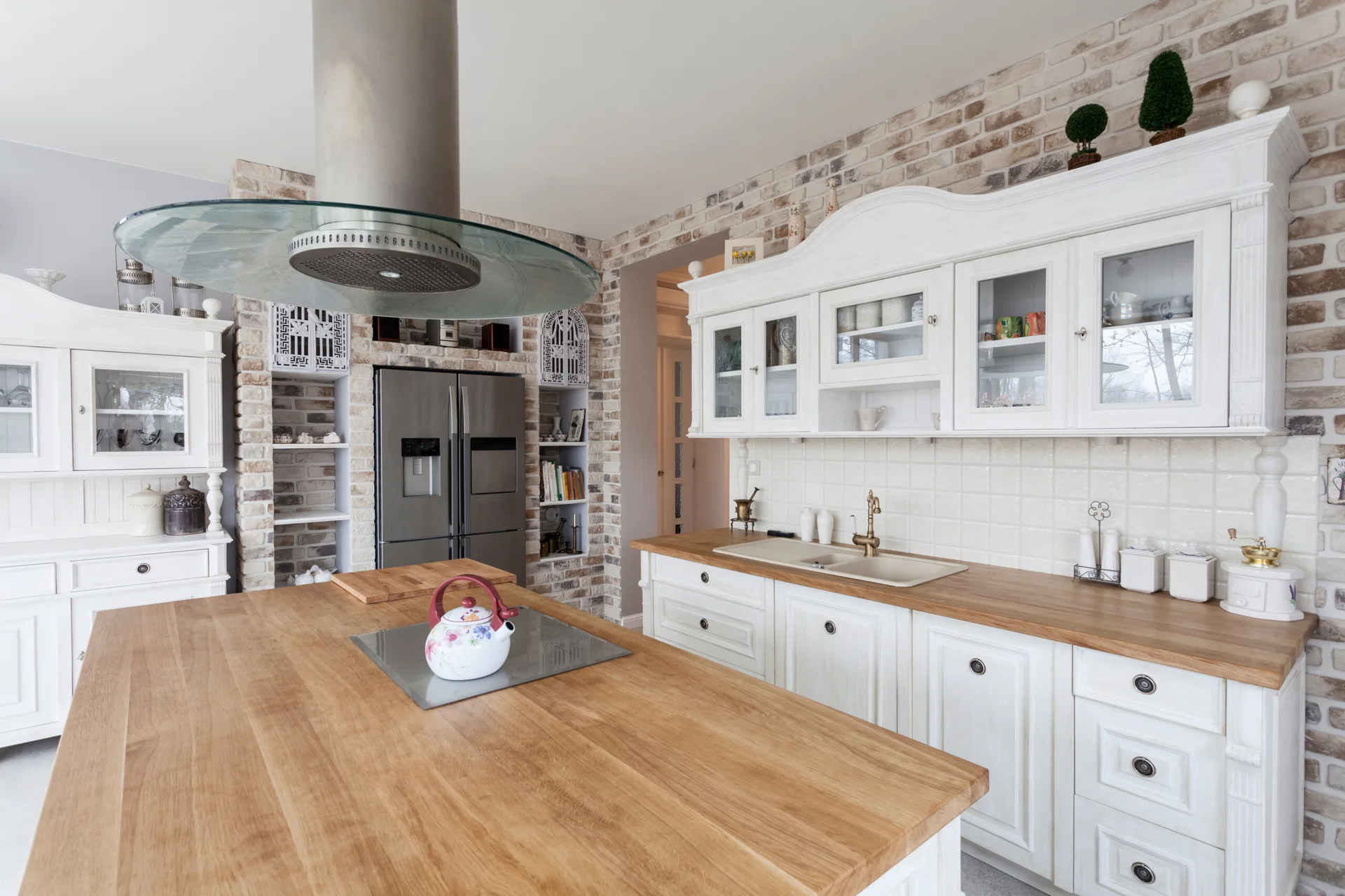 A farmhouse kitchen featuring white cabinetry, a large natural wood island with a kettle, and an exposed whitewashed brick wall.