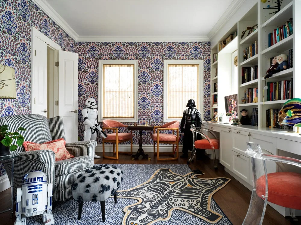 A colorful home study featuring floral patterned wallpaper, white built-in shelving, two coral chairs, a striped armchair, and pop culture figurines.