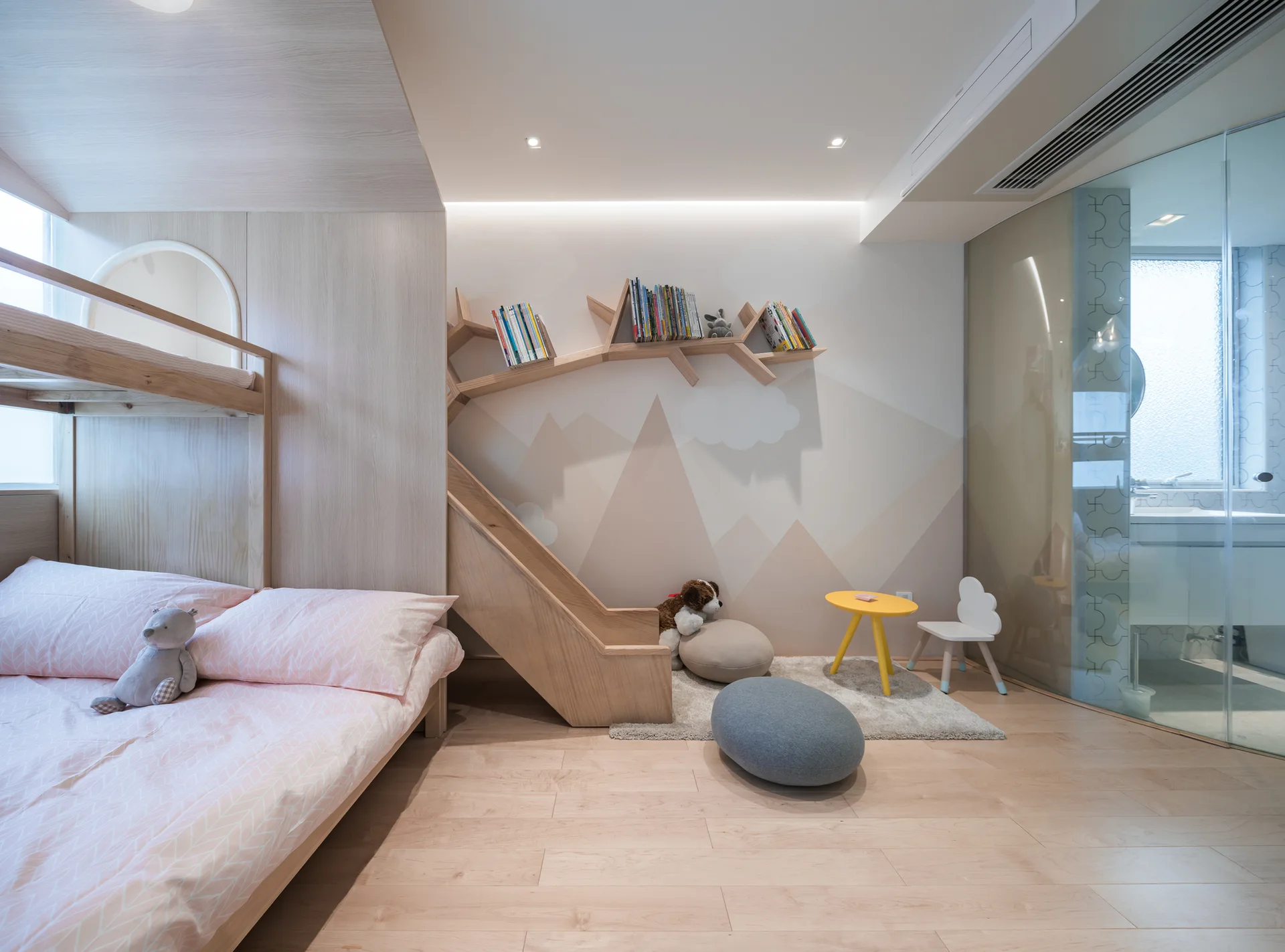 A light wood children's bedroom features a built-in bunk bed, a tree-shaped wall shelf against a mountain mural, and a small yellow play table.
