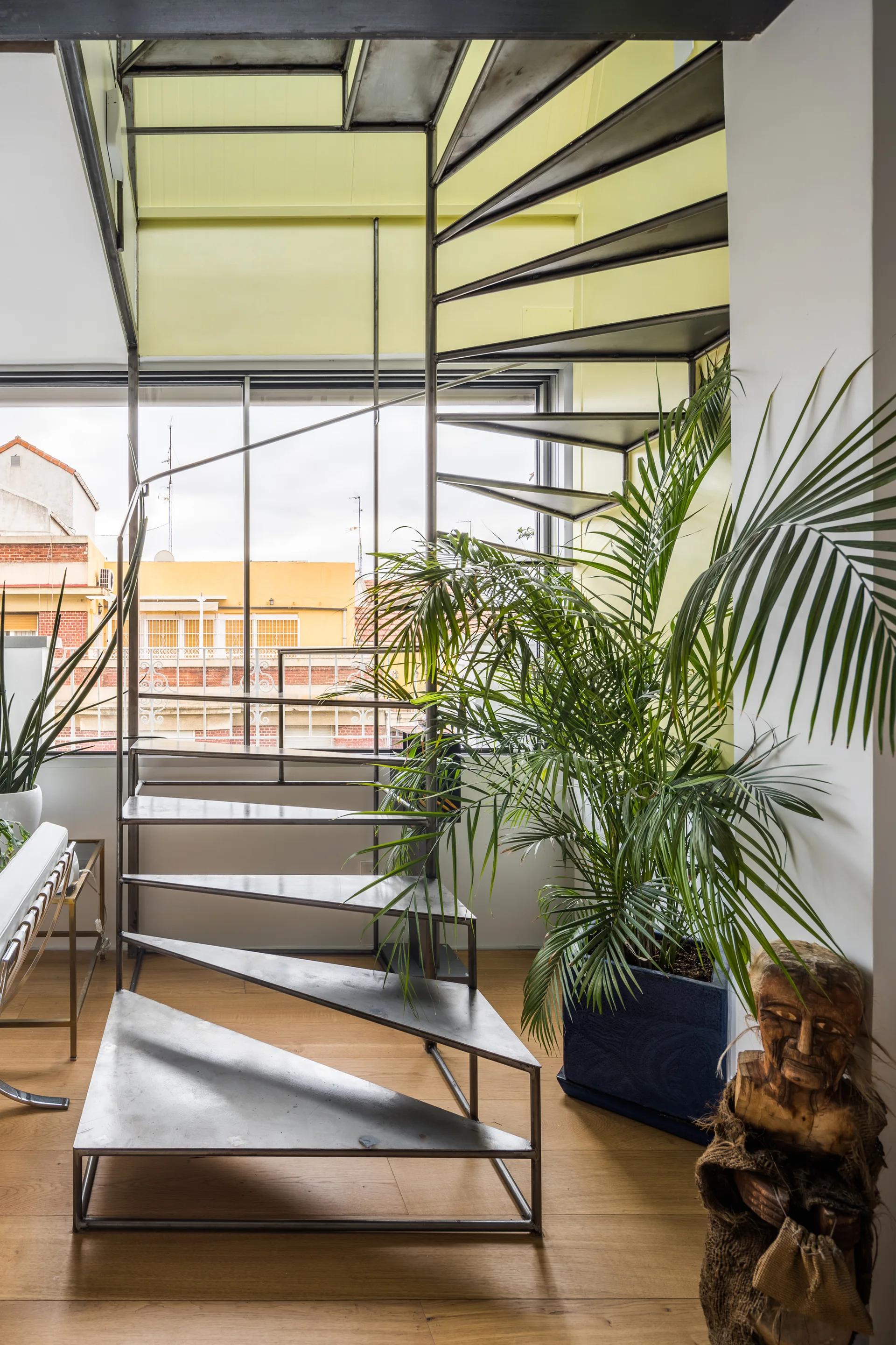 An industrial metal spiral staircase stands on light wood floors in front of a large window, next to a large potted palm plant.