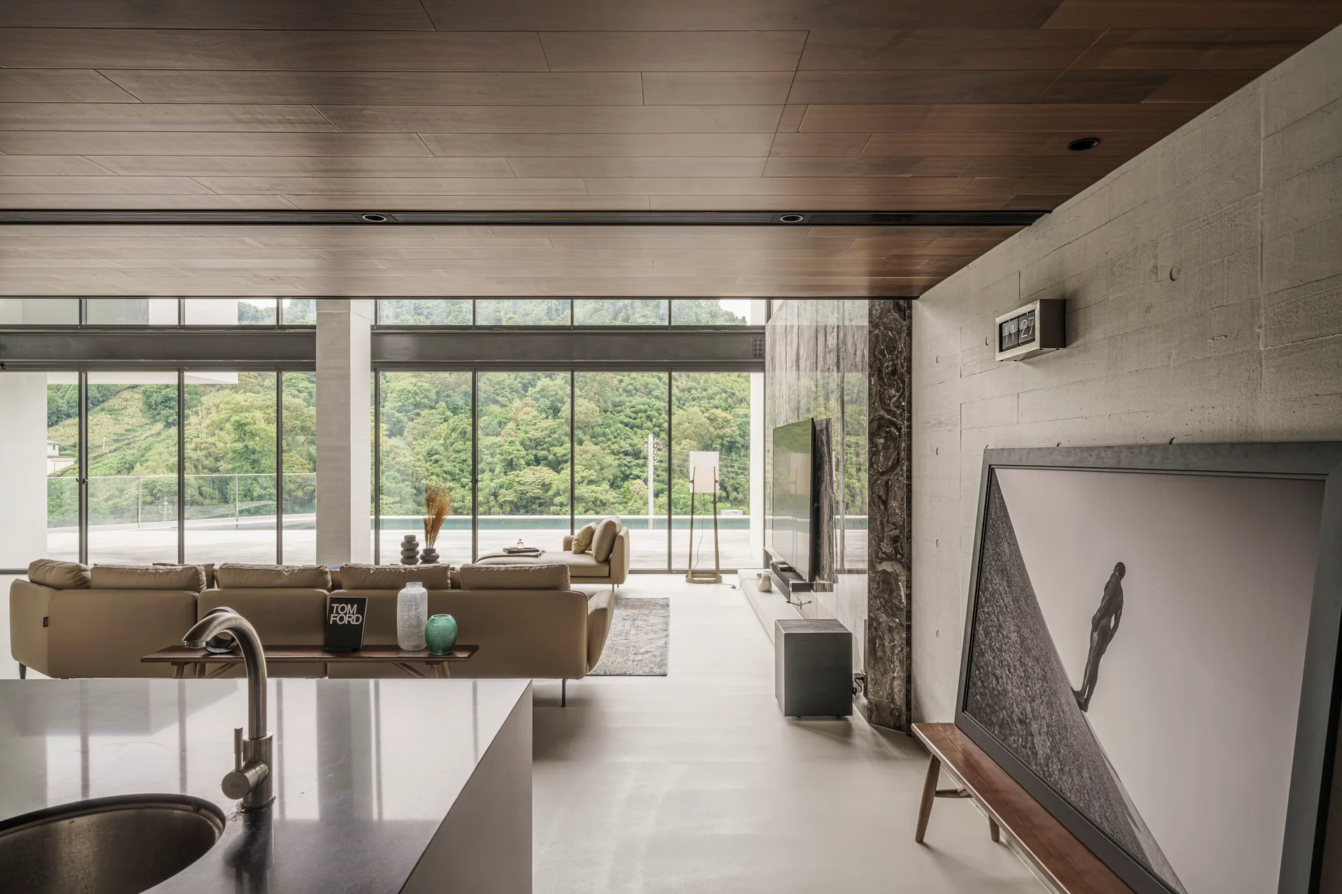 A modern living room with a tan leather sofa, board-formed concrete wall, wood ceiling, and floor-to-ceiling windows showing an outdoor pool.