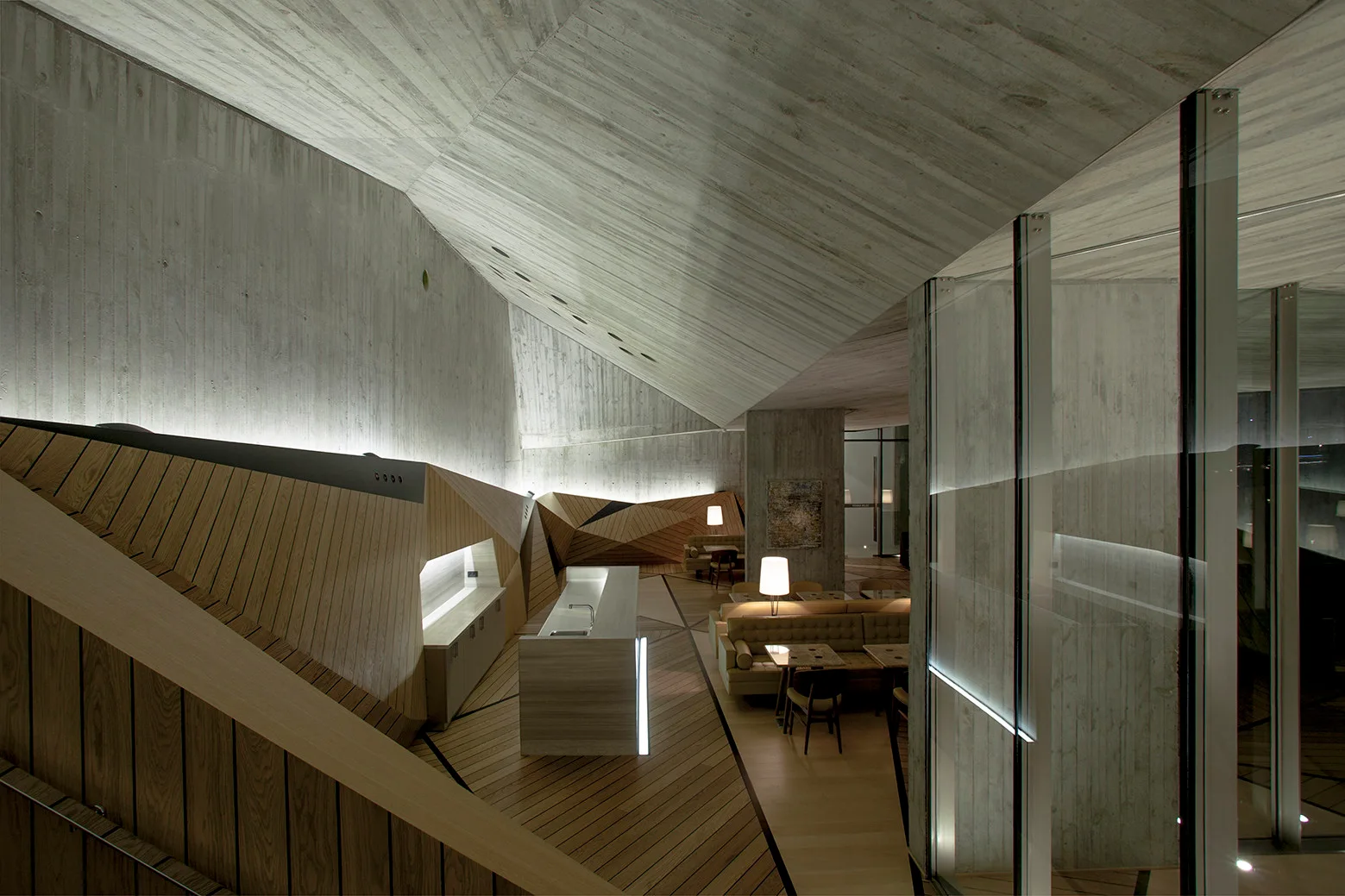 A modern architectural interior featuring a dramatic sloped concrete ceiling, geometric wood wall paneling, a wooden floor, and a low seating area.