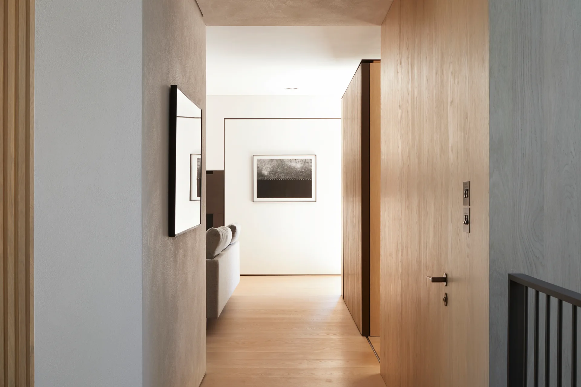 A minimalist hallway with light wood flooring, a flush wooden door, and slatted paneling leading to a bright living room with a framed art piece.