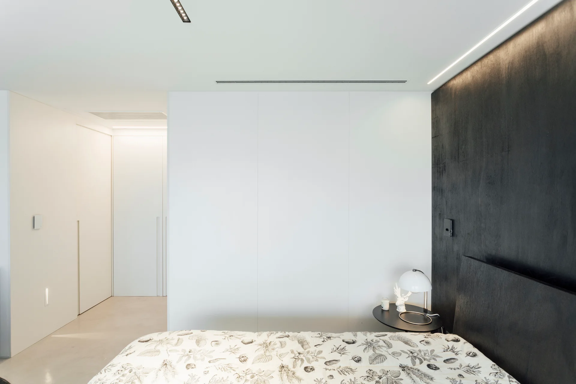 A minimalist bedroom featuring botanical print bedding, a small round black side table with a dome lamp, and a matte black accent wall panel.