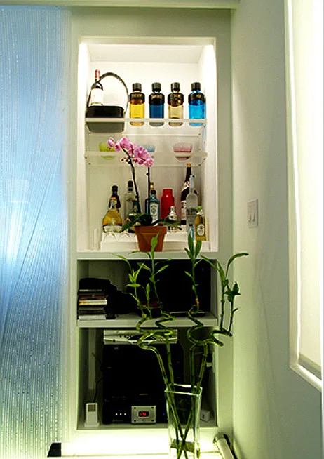 A built-in white bar niche holds liquor bottles, glassware, and an orchid above lower shelves with audio equipment.