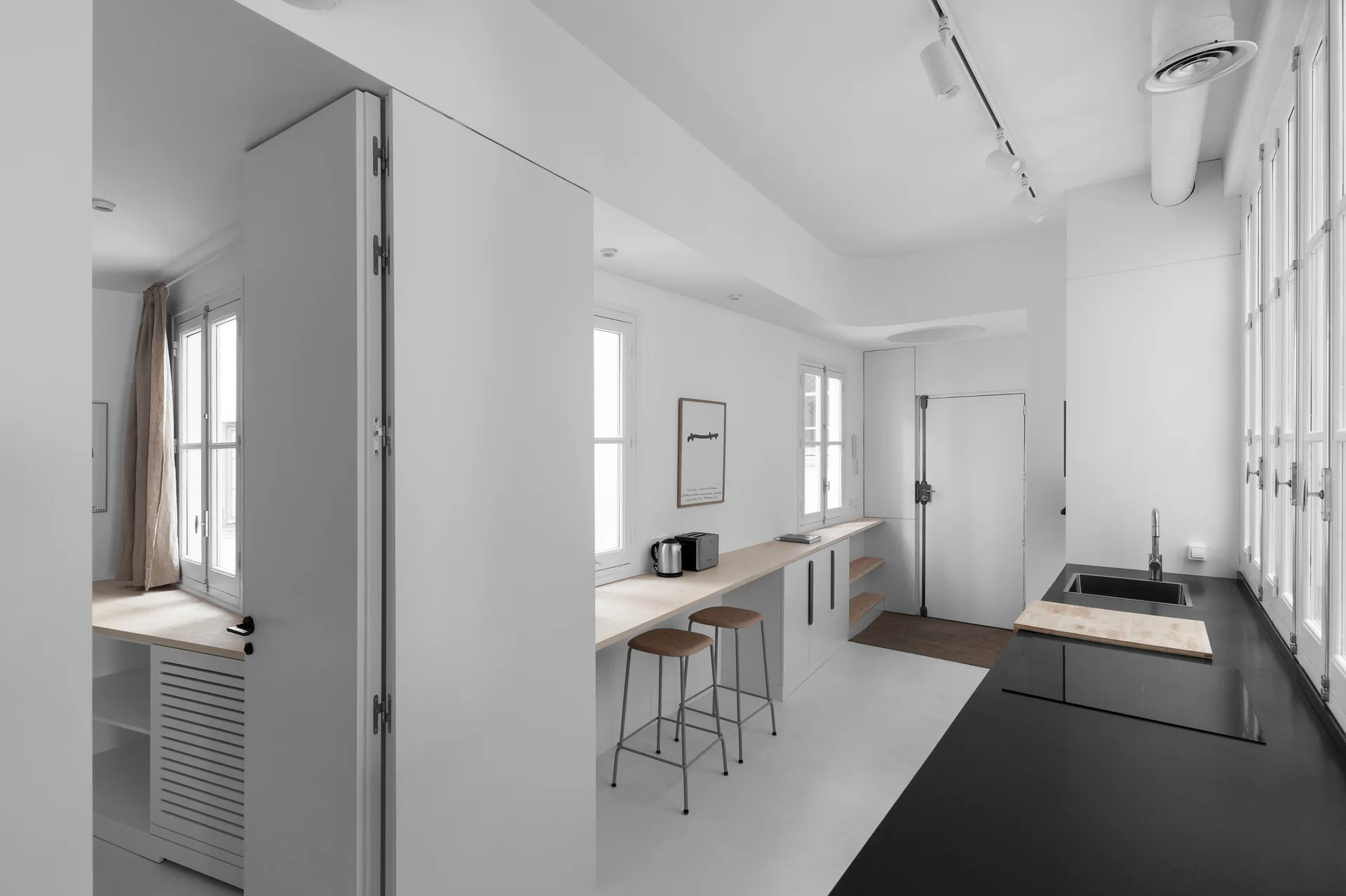 A minimalist white kitchen features a long light wood bar counter with two wooden stools and a contrasting black countertop with a sink.