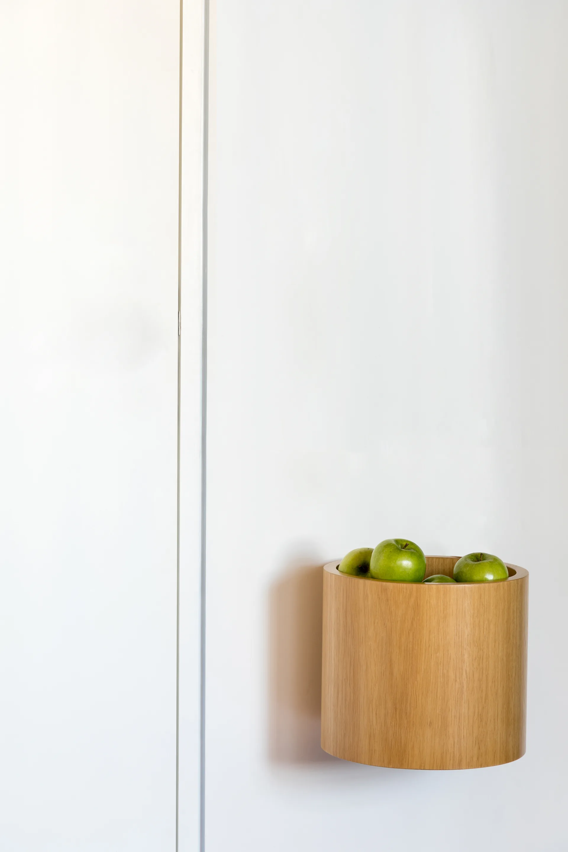 A cylindrical natural wood container holding green apples is mounted on a smooth white wall.