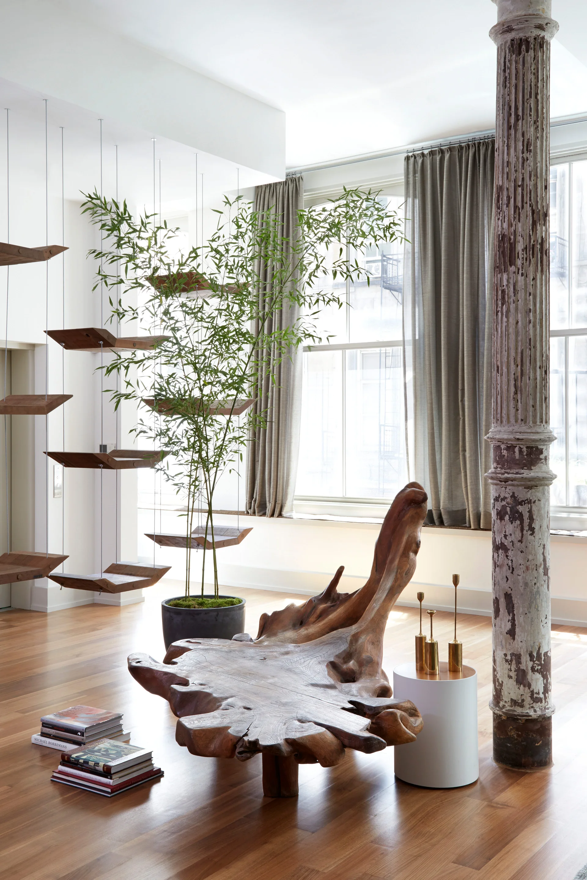 An open loft living space features a sculptural live-edge wooden bench, a weathered structural column, and suspended wood shelving near a large window.