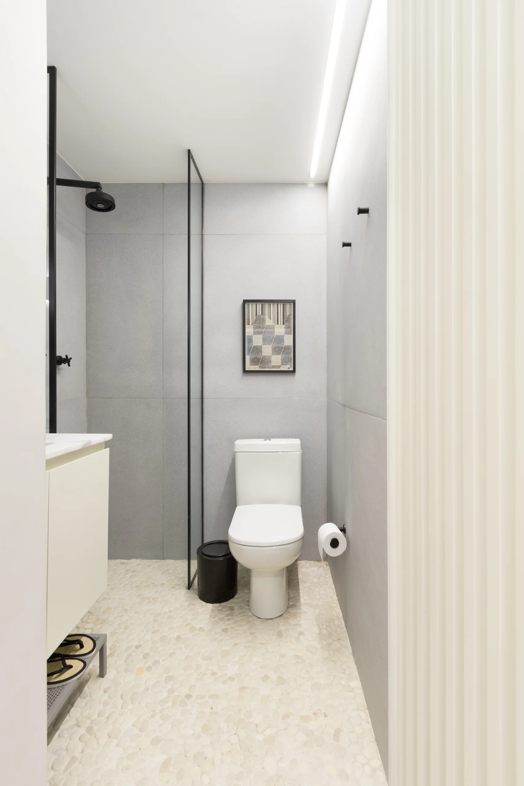 A minimalist bathroom featuring matte grey wall tiles, a white ceramic toilet, a glass shower partition, and a textured pebble floor.
