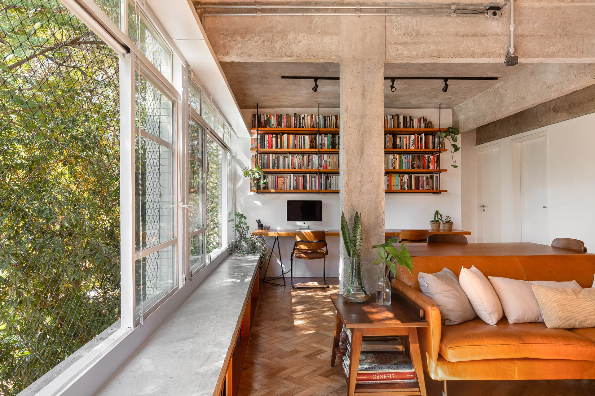 An industrial home office with a tan leather sofa, herringbone wood floors, and a desk arranged near an exposed concrete column.