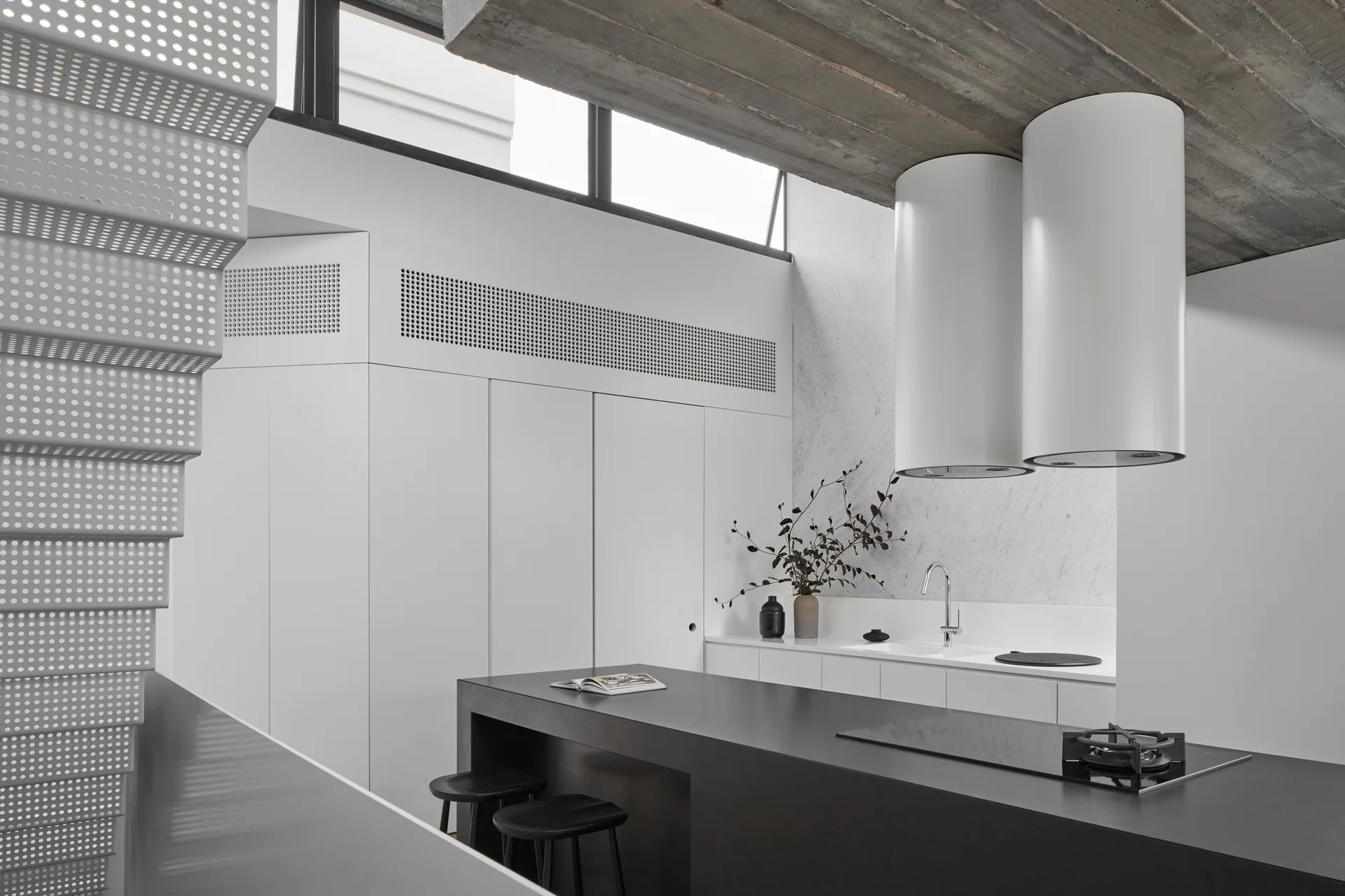 A minimalist modern kitchen with a dark central island, dual white cylindrical range hoods, and an exposed raw concrete ceiling.