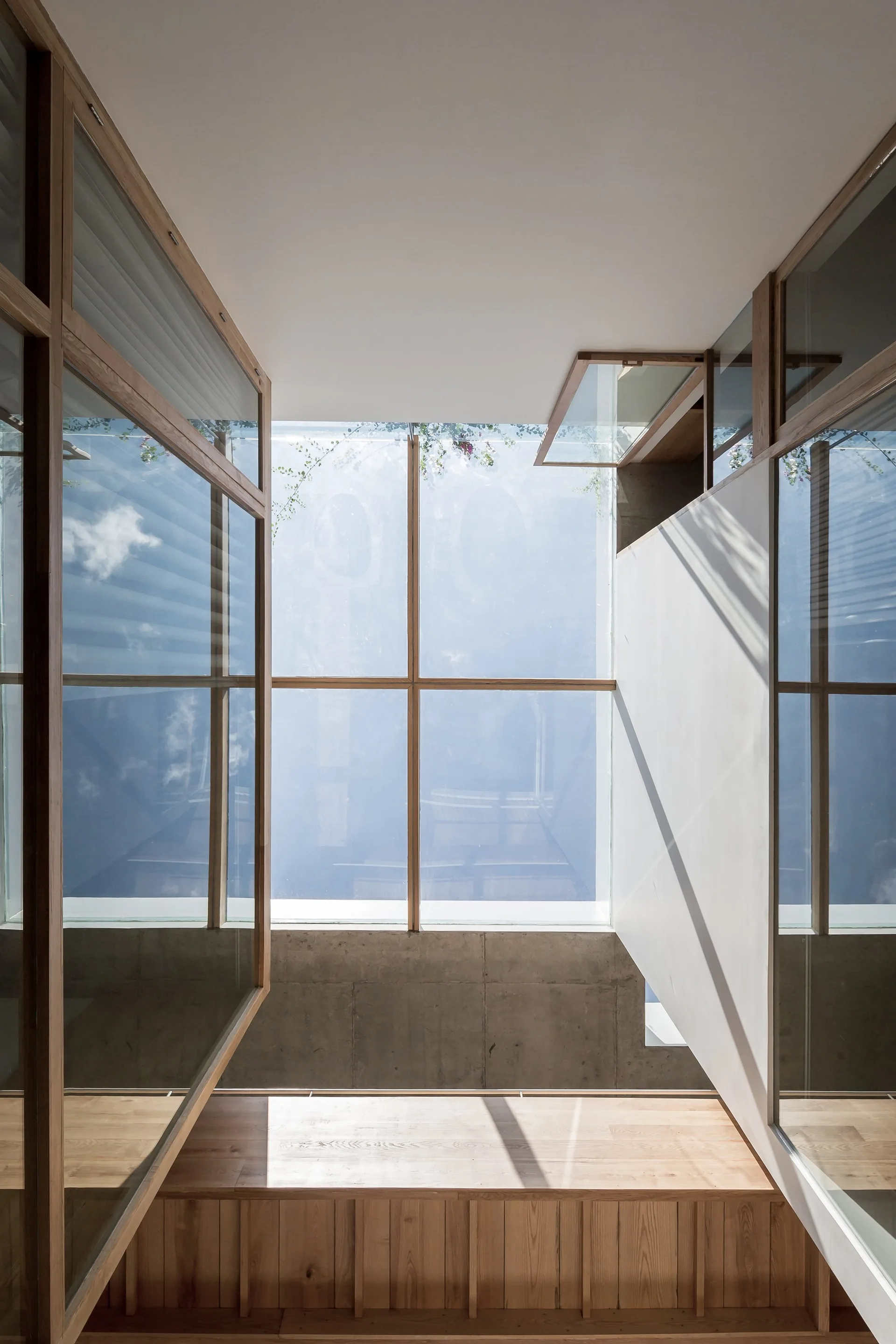 A vertical view looking up at a large glass skylight with light wood framing, surrounded by concrete walls and a wooden built-in bench below.