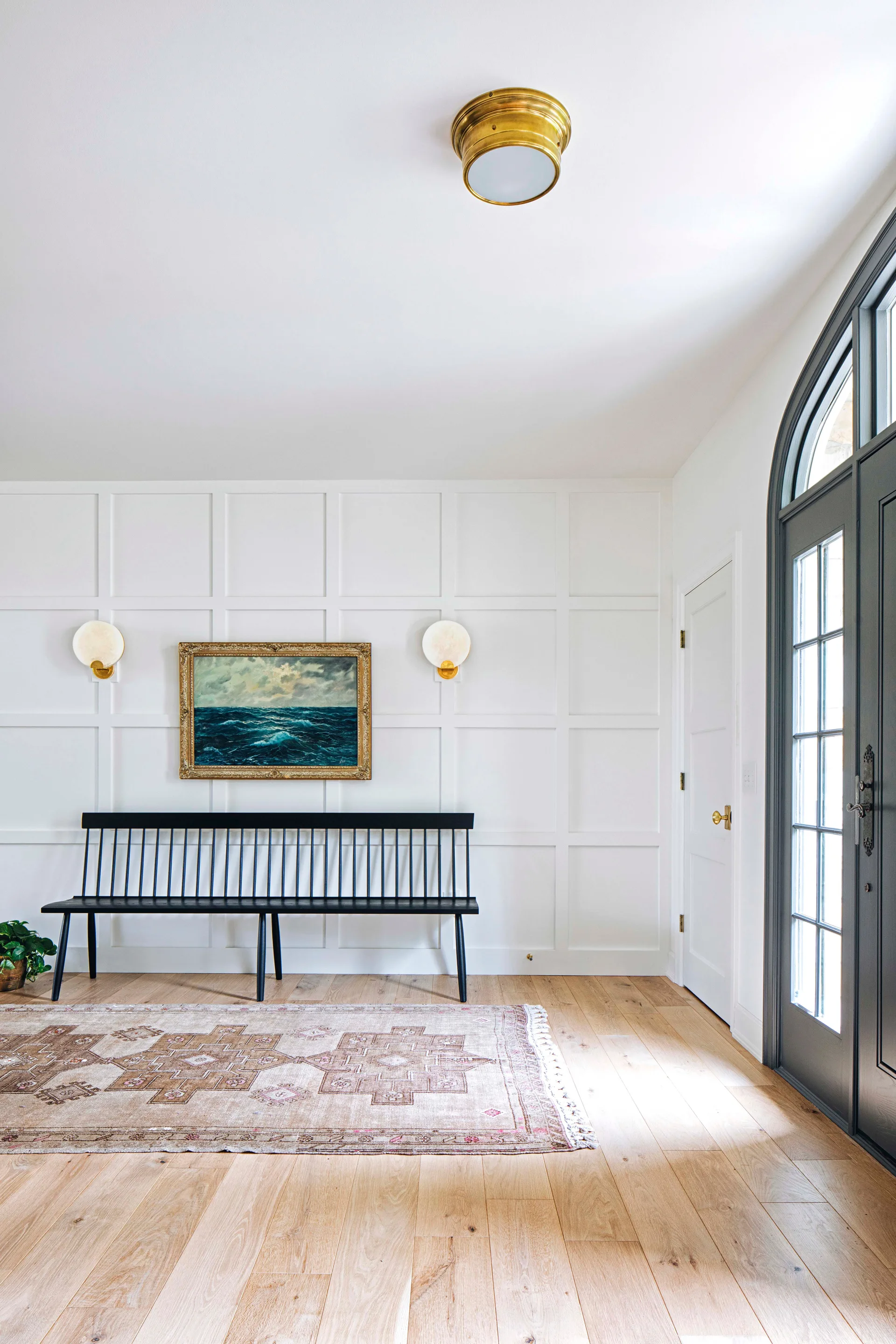 A black spindle bench sits against a white grid-paneled wall in an entryway next to a dark grey arched door and a patterned vintage rug.