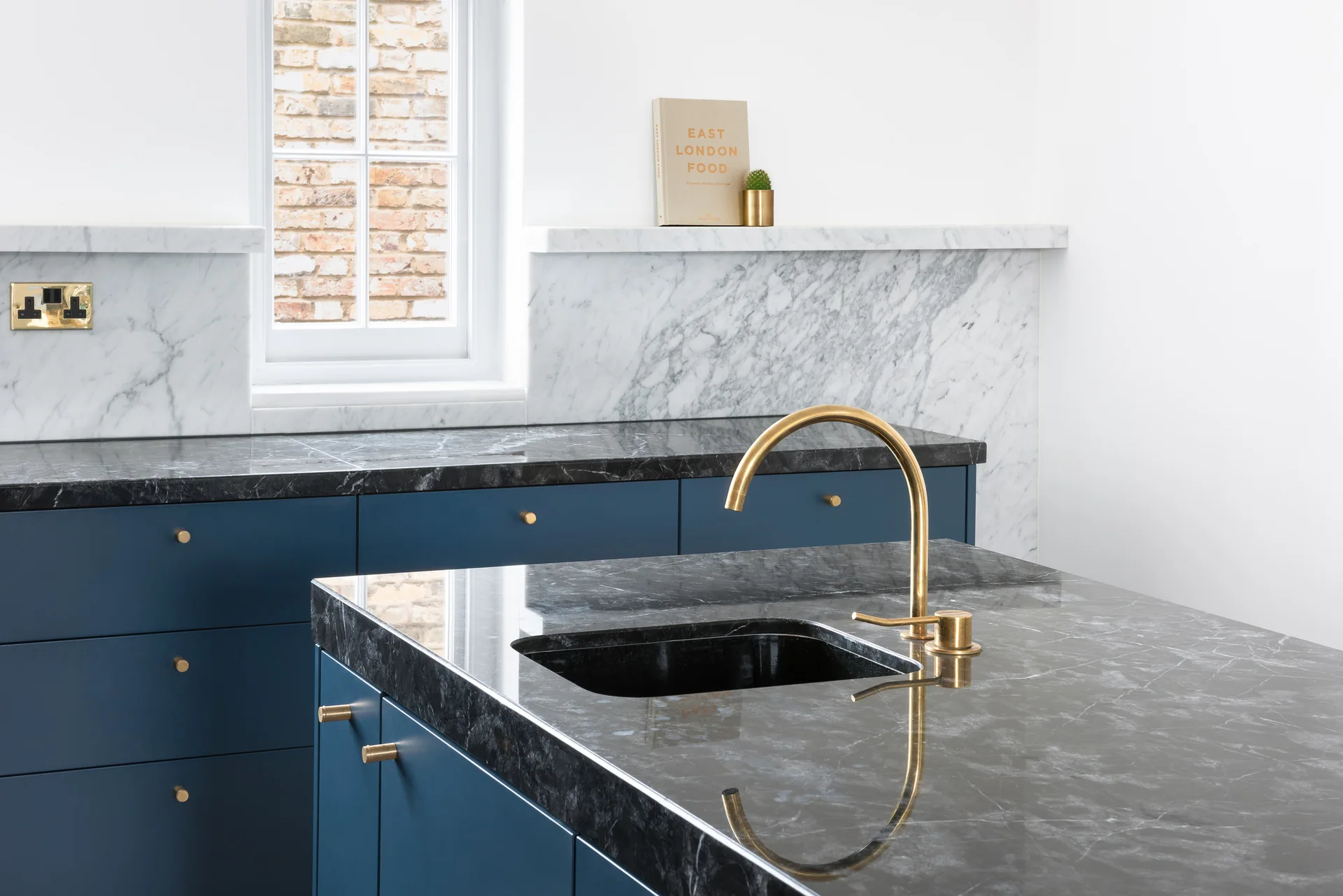 A modern kitchen featuring navy blue cabinets with brass handles, a black marble island countertop with a brass faucet, and a white marble backsplash.