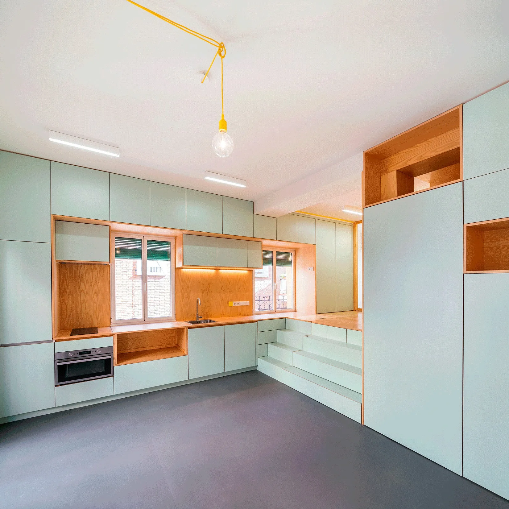 A minimalist kitchen features pale green cabinetry with integrated wood shelving, built-in stairs, and a single yellow pendant light.